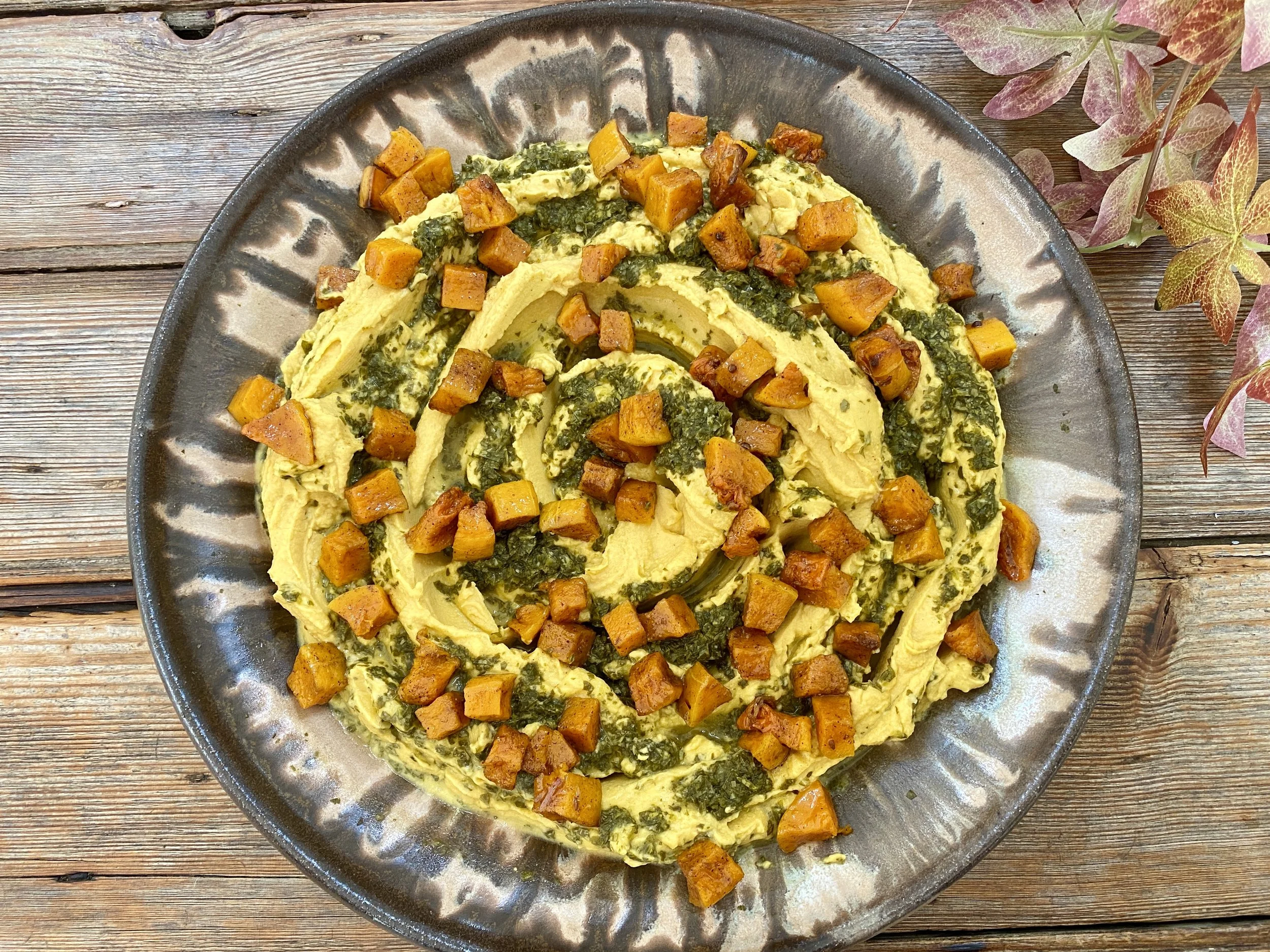 A plate of mashed yellow potatoes topped with roasted sweet potatoes and greens, served on a rustic wooden table with some pink and green leaves nearby.