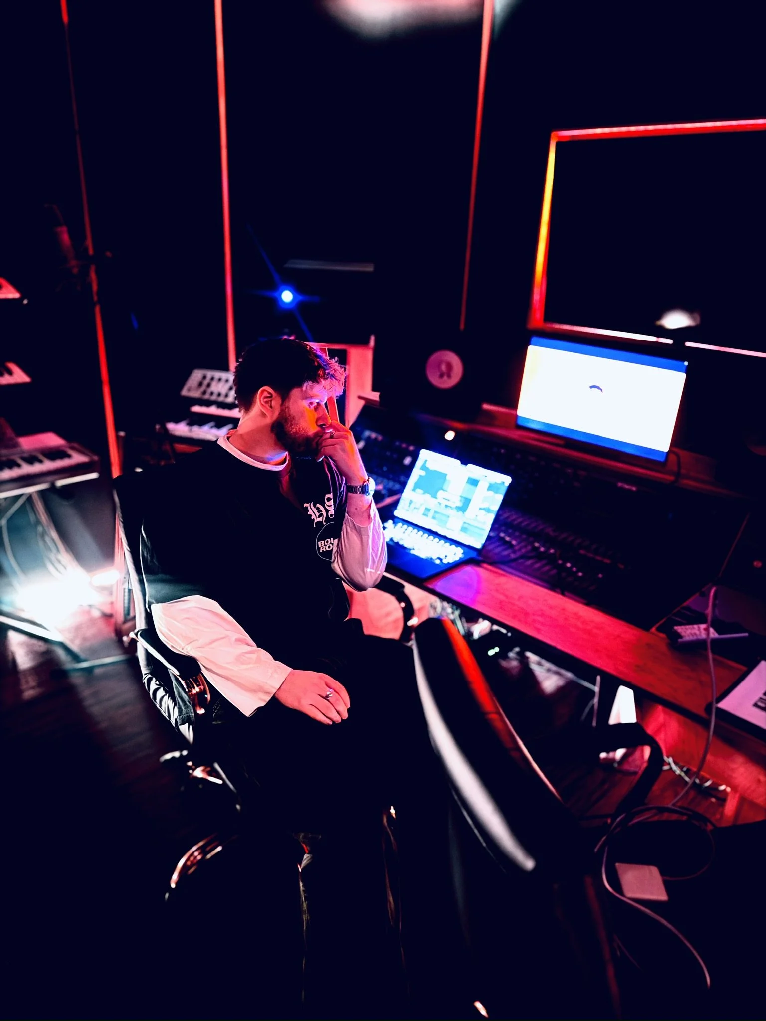 A man sitting in a recording studio with music production equipment, including a keyboard, computer monitors, and a mix console, illuminated by black and red lighting.