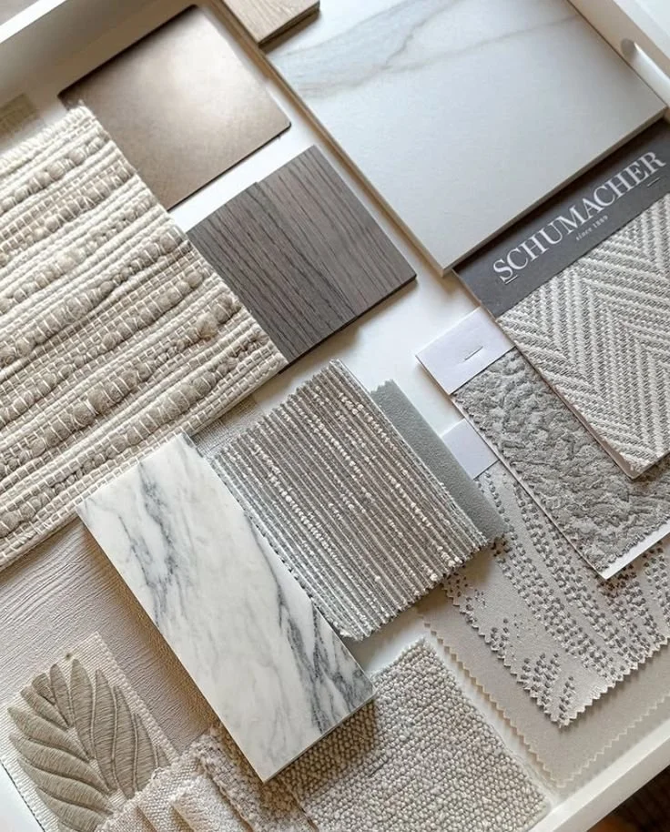 Collection of various fabric, tile, and material samples in neutral tones, including fabric textures, marble, and patterned surfaces, arranged on a white surface.