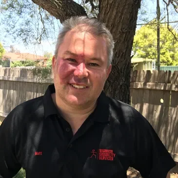 A man with short gray hair smiling outdoors in front of a tree and wooden fence, wearing a black polo shirt with red text and logos.