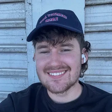A young man with short, curly hair and a beard, smiling. He's wearing a black baseball cap and a black shirt, and has white wireless earbuds in his ears. The background features a gray garage door.