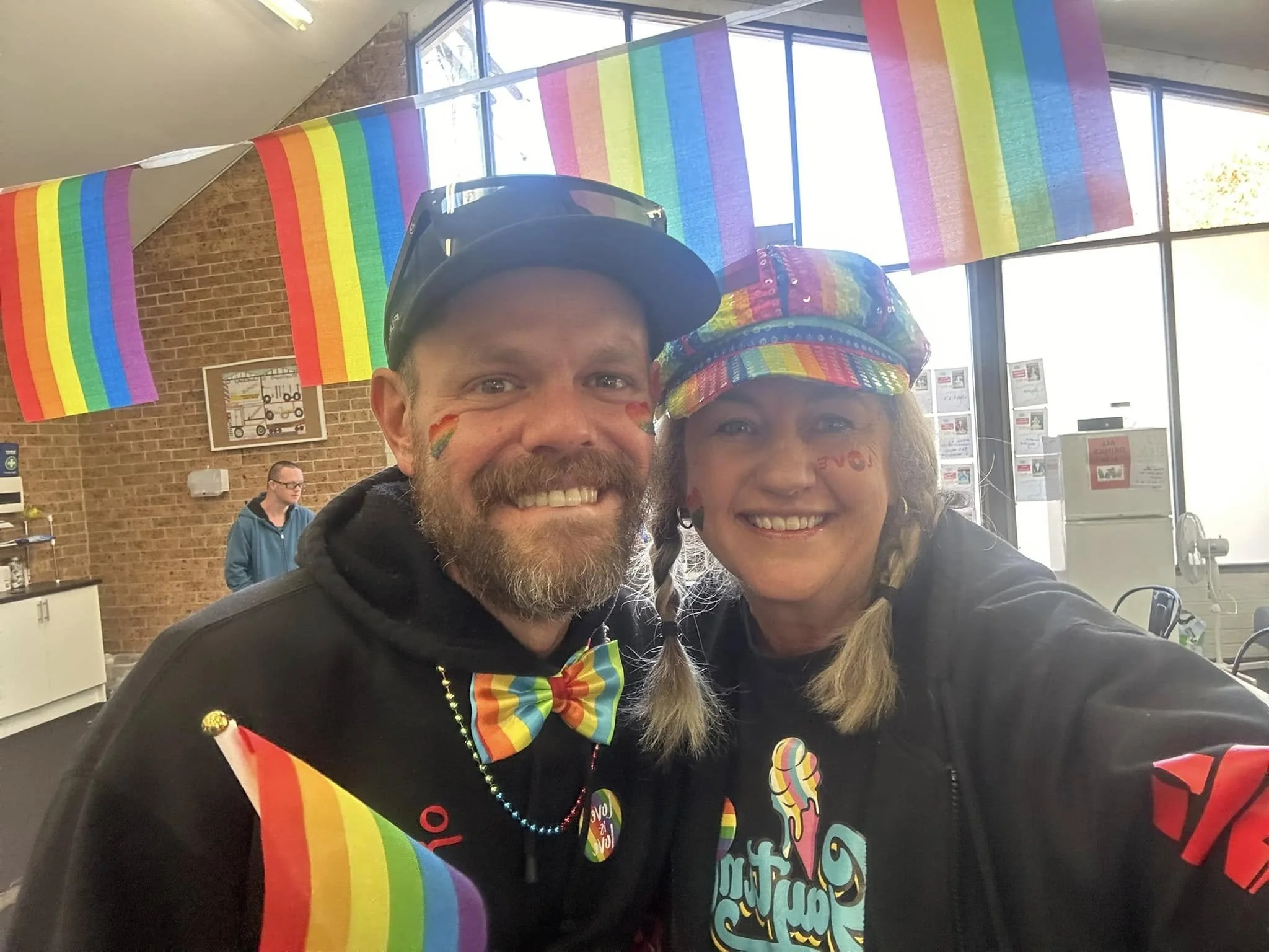 Two people smiling at a pride celebration, decorated with rainbow flags and colorful accessories, in a room with large windows and brick walls.