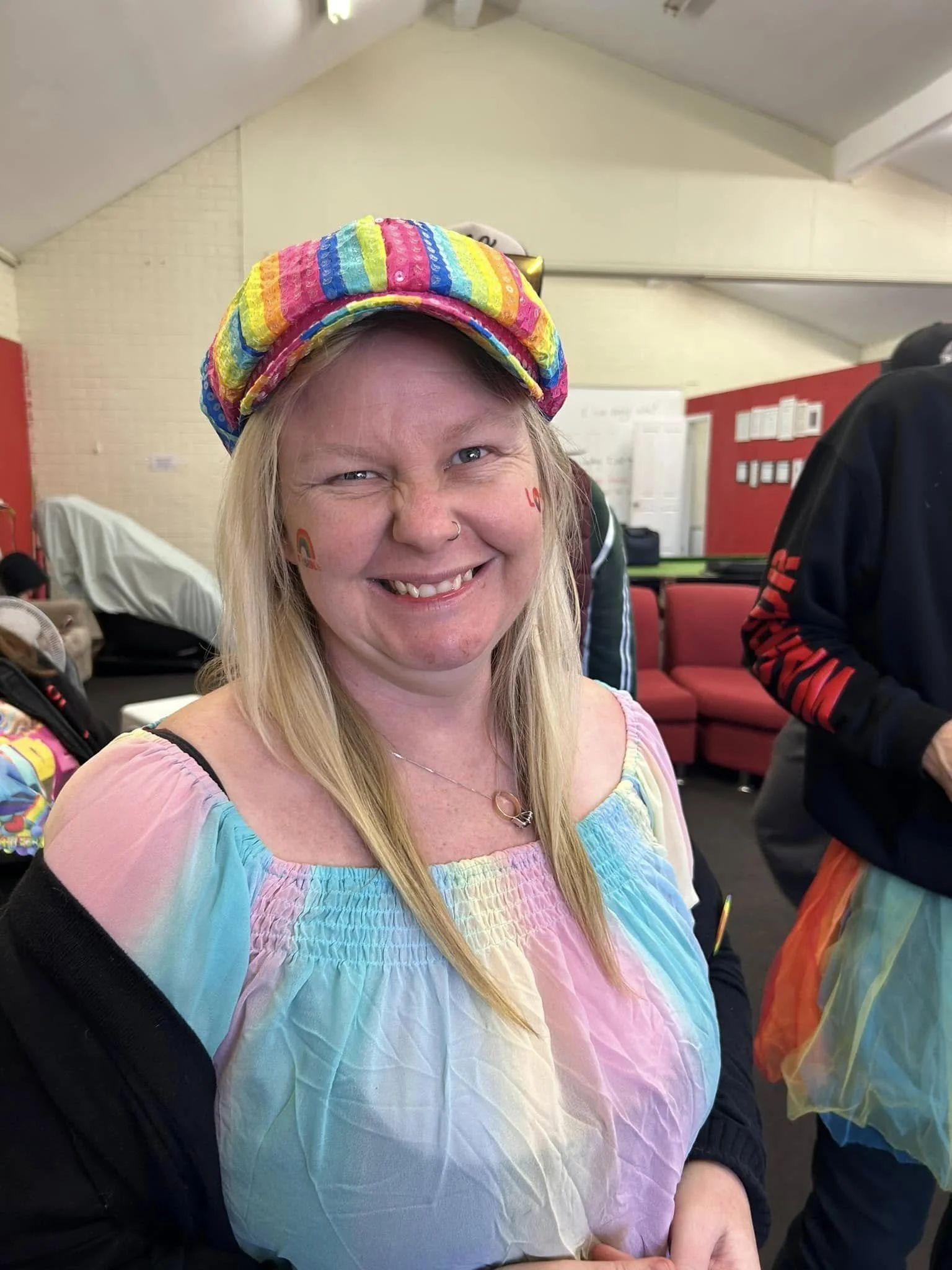 A woman smiling at the camera, wearing a rainbow-colored hat and a pastel rainbow tie-dye shirt, with small rainbow tattoos on her cheeks, in an indoor setting with red and white chairs and whiteboards in the background.