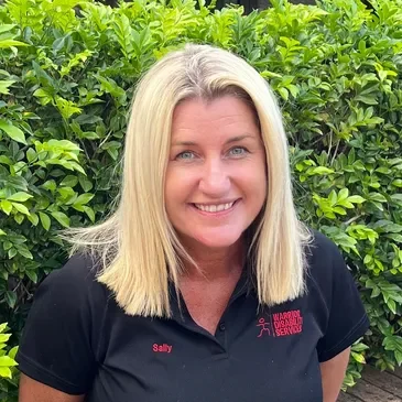 Smiling woman with blonde hair wearing a black polo shirt with red logo and name tag, standing in front of green bushes.