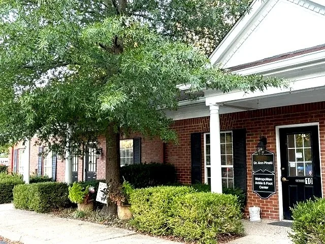 A brick building with black shutters, a black door, and a sign that says 'Dr. Ann Prasad, Podiatrist, and Foot & Ankle Center.' A large green tree and neatly trimmed bushes are in front, along with a sidewalk.