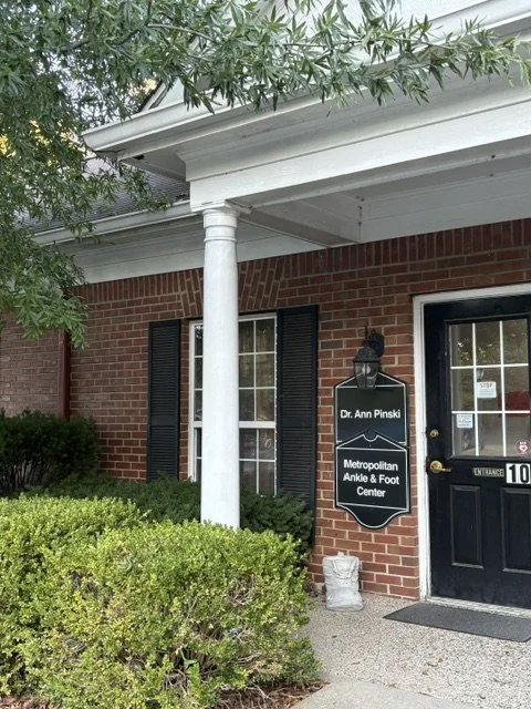 Sign on brick building reading 'Dr. Ann Pinski, Metropolitan Ankle & Foot Center' with black door, window with black shutters, and green bushes in front.