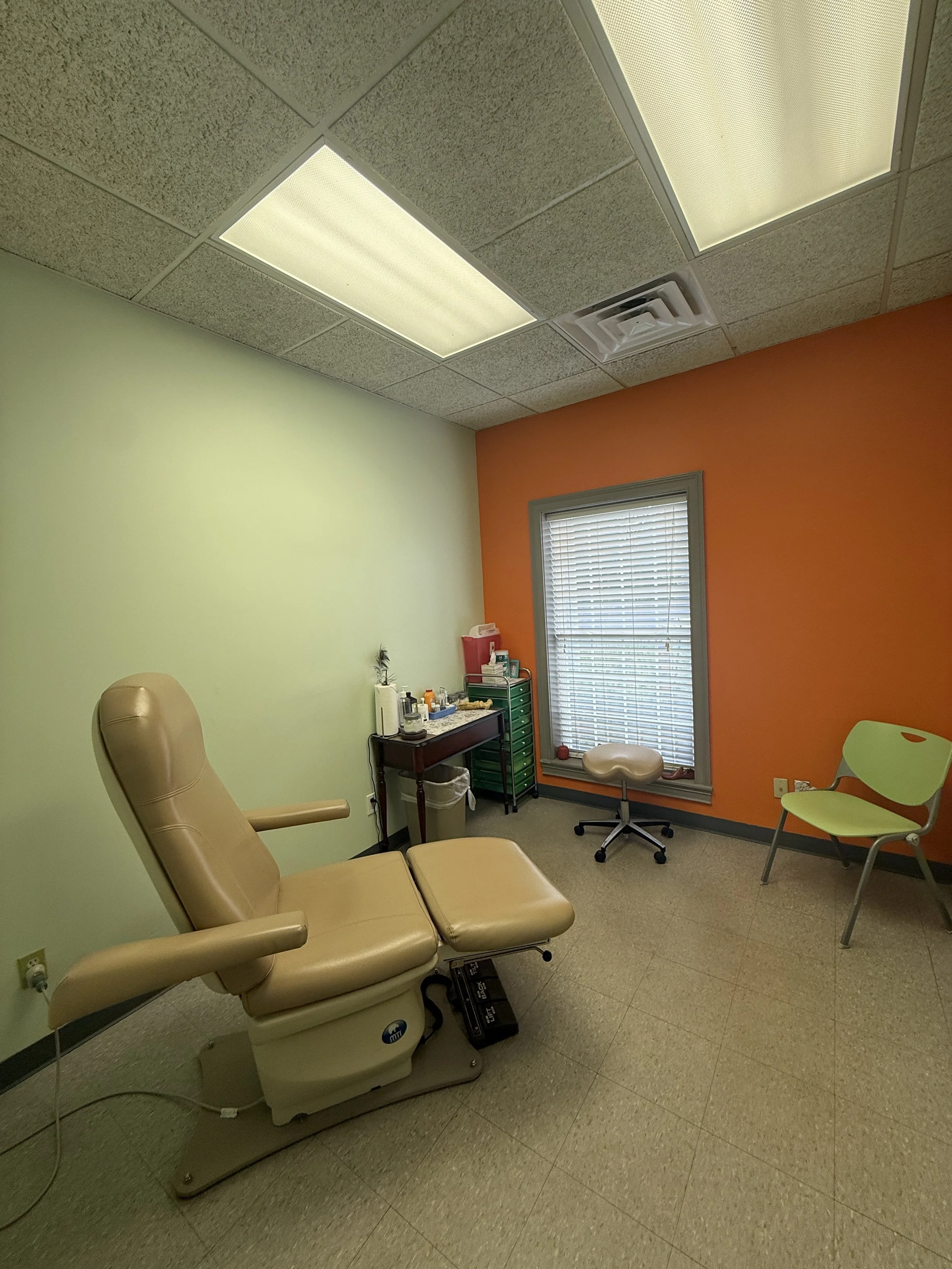 Medical examination room with a beige reclining examination chair, a small desk with medical supplies, a green cabinet, two chairs (one green, one beige), and a window with blinds. The ceiling has fluorescent lights and an air vent.