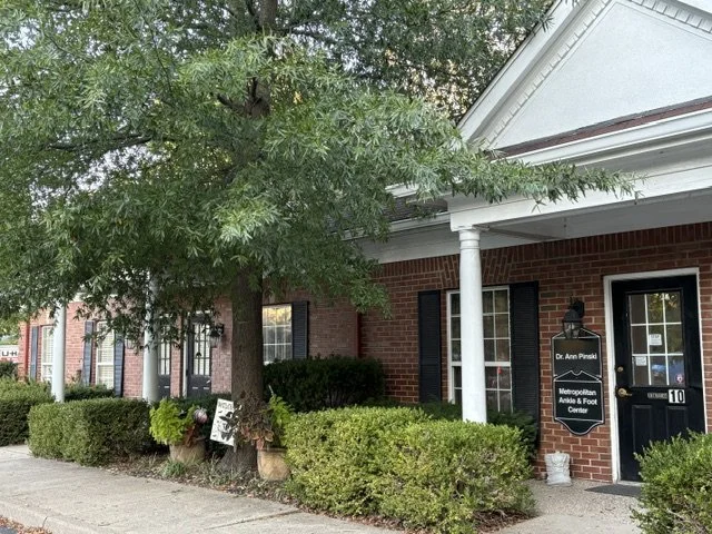 Exterior view of a brick building with a black door, white columns, and a sign for Dr. Ann Pinski, featuring bushes, a tree, and sidewalk.
