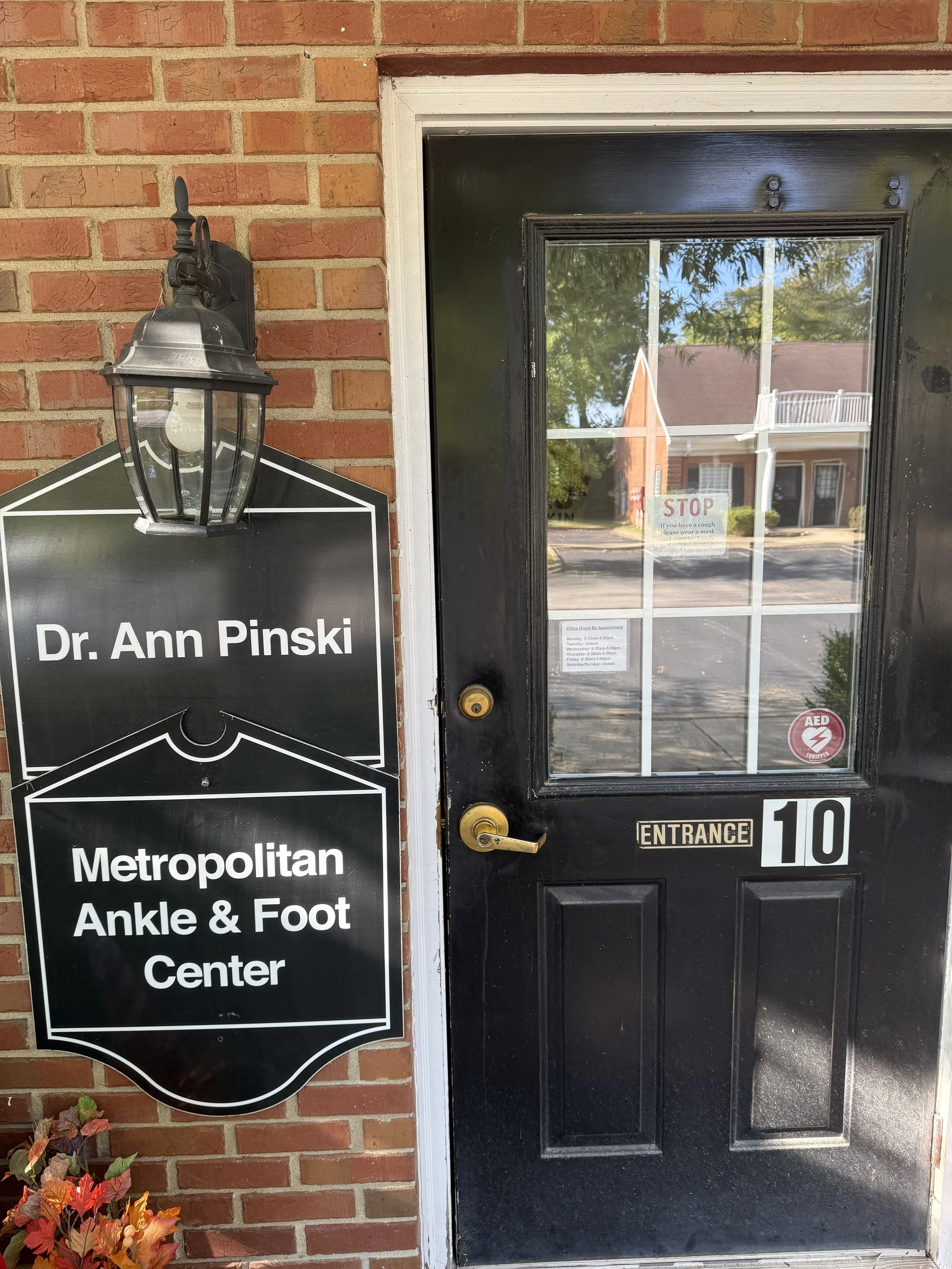 Black door with gold handle and lock, glass window, sign on the glass saying 'STOP if you have a cough, please wear a mask, and sanitize before entering,' and a sticker showing an AED. To the left, a black lantern-style light fixture and a black sign with white text reading 'Dr. Ann Pinski, Metropolitan Ankle & Foot Center' is mounted on a brick wall with autumn leaves at the bottom left.
