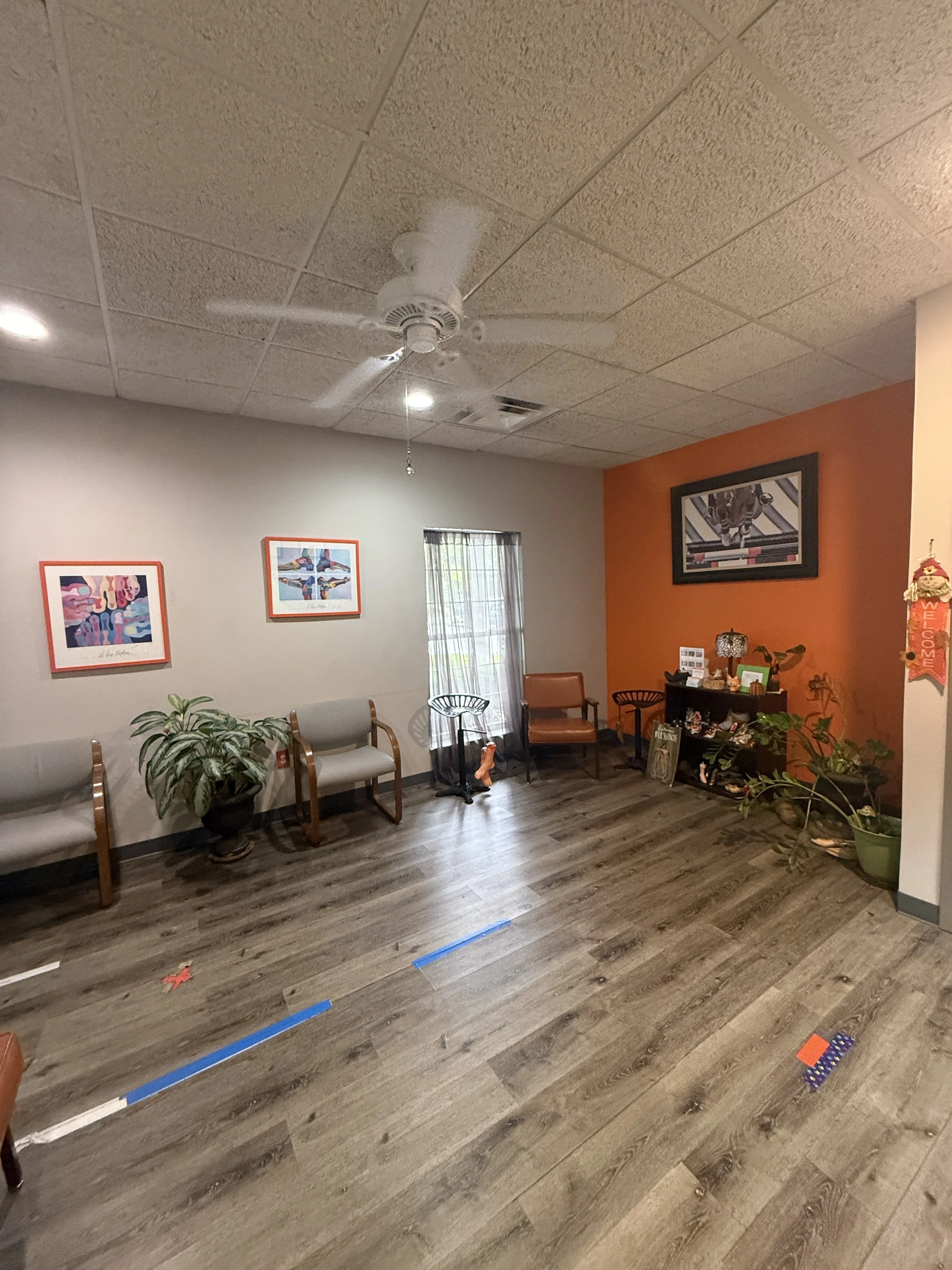 An indoor room with a gray wall, orange accent wall, wooden floor, several chairs, plants, and framed artwork on the walls. Ceiling lights and a ceiling fan are also visible.