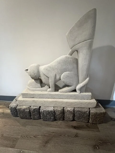 Stone sculpture of a cat with a modern abstract high heel behind it, placed on a stone base on a wooden floor