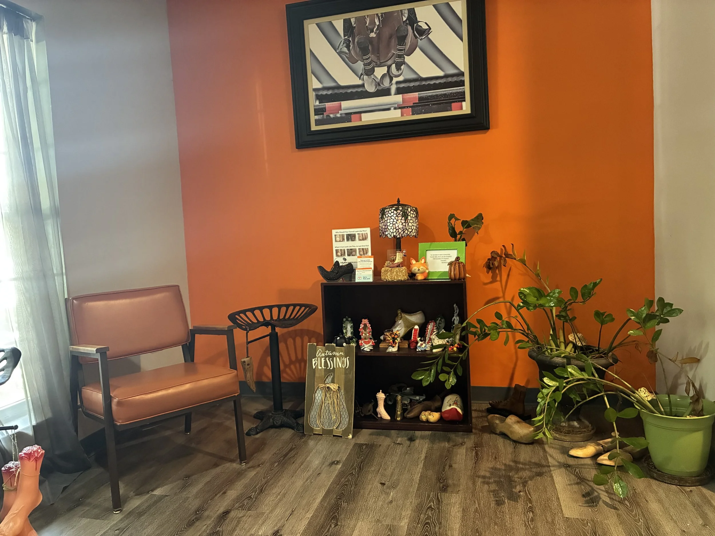 Interior of a room with orange and cream walls, decorated with a framed picture of a horse jumping over a hurdle, a table with small decorative items, a pink chair, a black chair, and several potted plants. There are shoes and a sign that says "Autumn blessings" on the table.