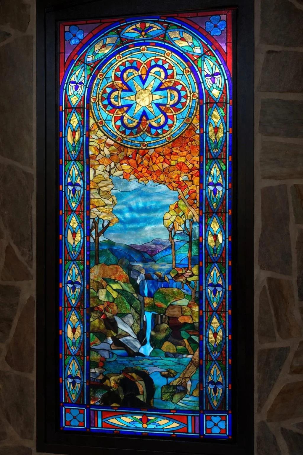A stained glass window depicting a landscape with a waterfall, trees, mountains, and a blue sky, surrounded by intricate geometric patterns and symbols with vibrant colors.
