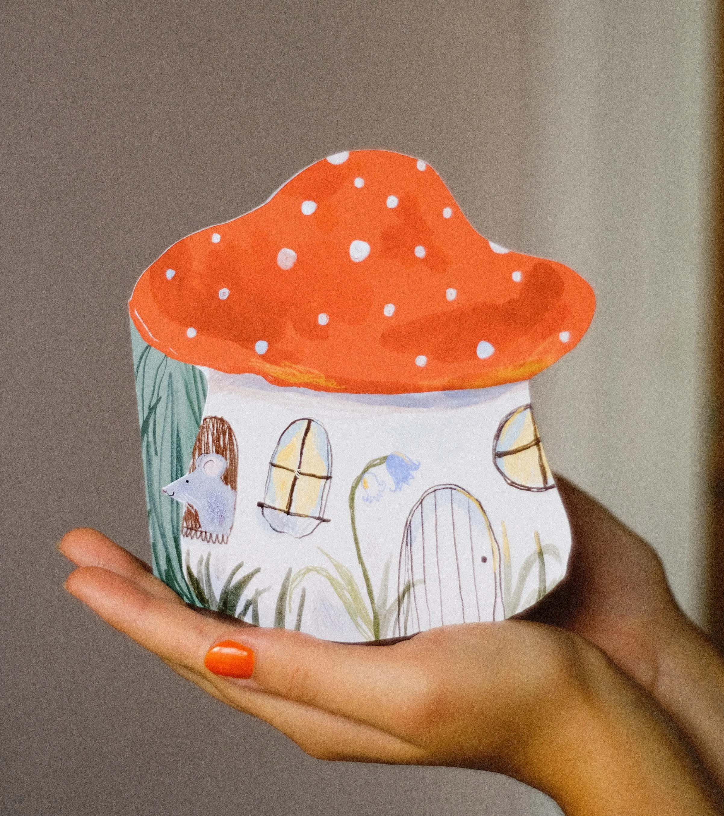 A handmade card in the shape of a toadstool. This illustration is cute and colourful, where the toadstool poses as a house for small mice characters, one of them is sticking their head out a window made of branches.