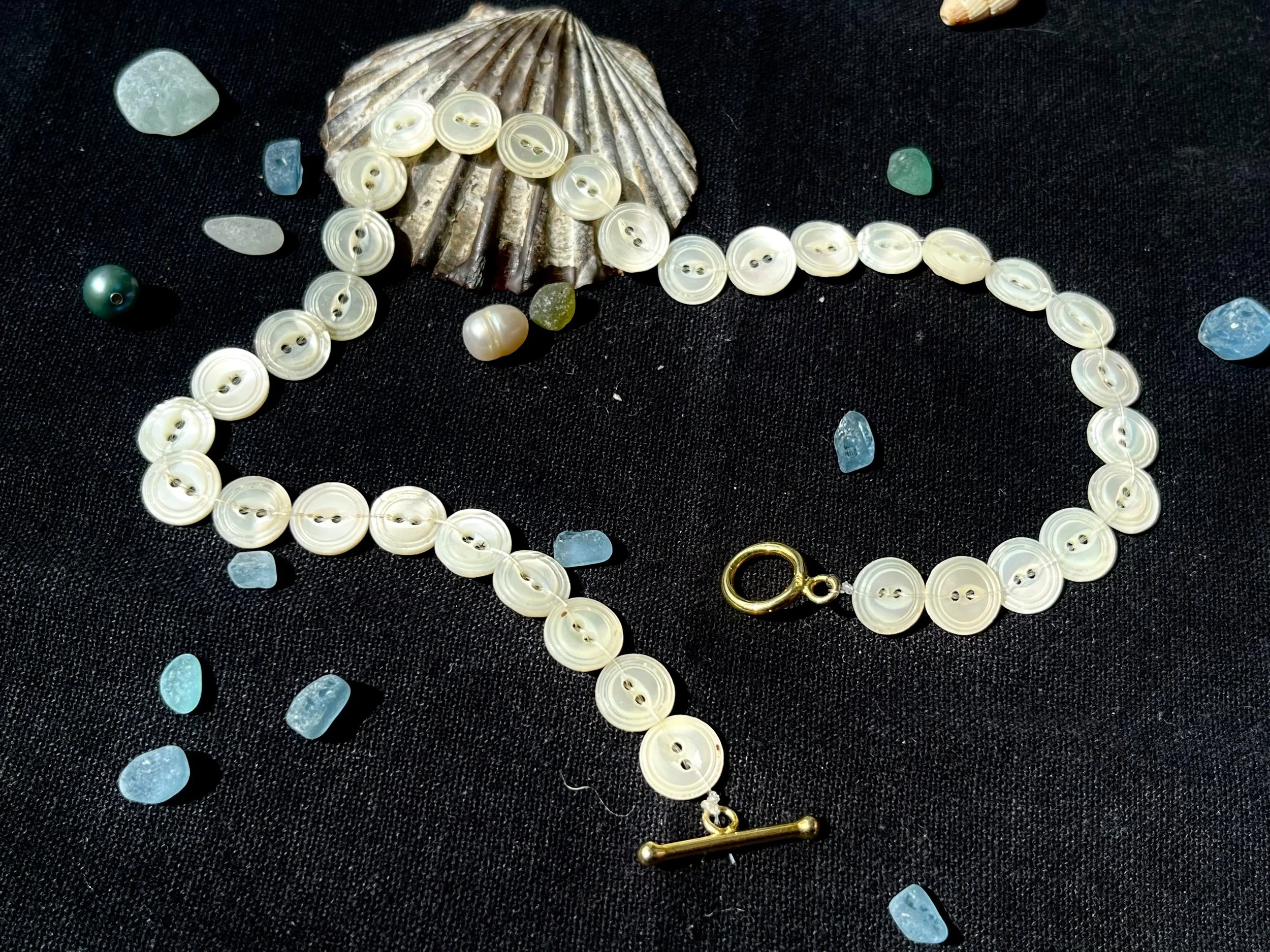 An assortment of seashells, pearls, and colorful gemstones arranged on a black fabric background, including a large scallop shell in the center.