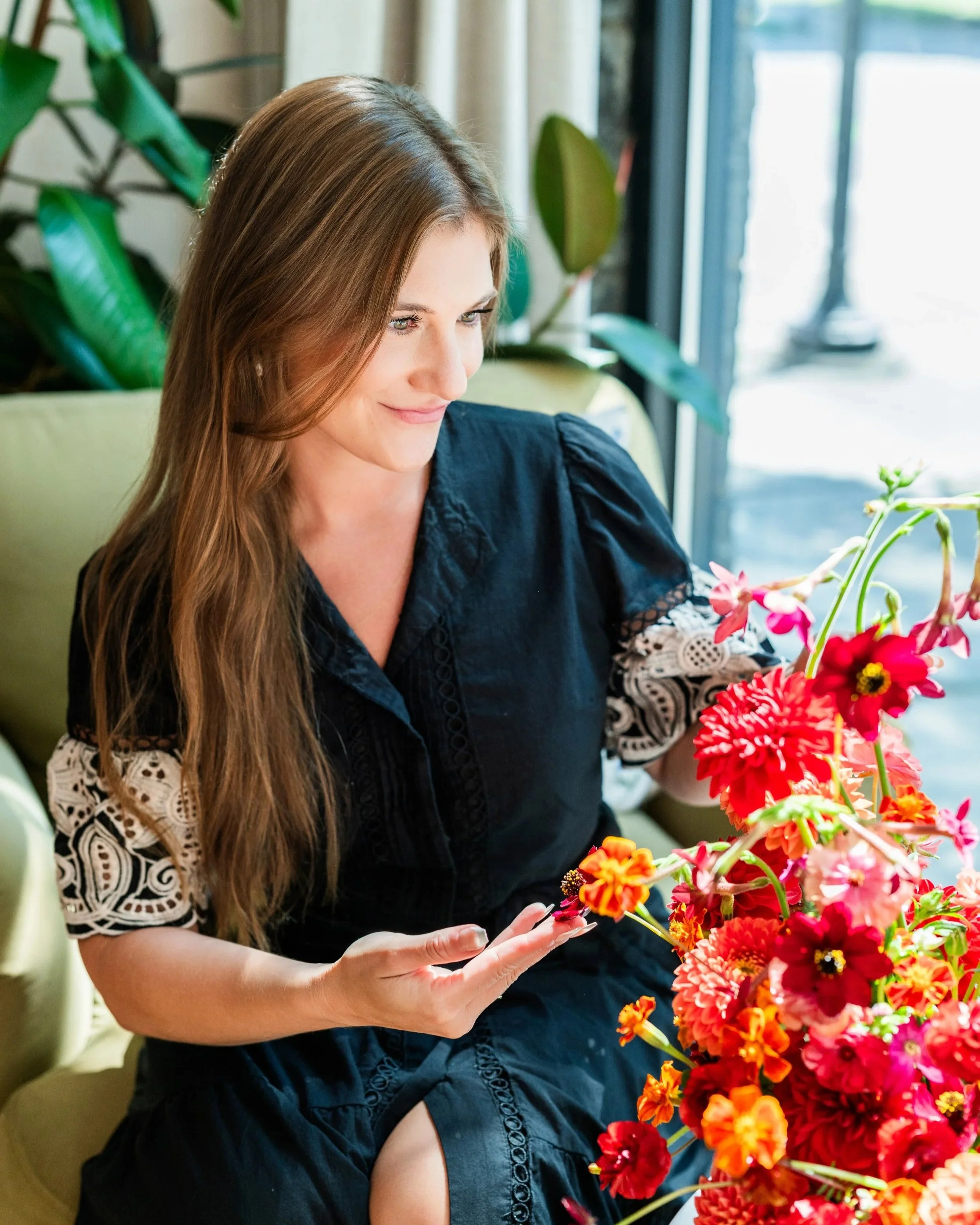 A woman with long brown hair and green eyes arranging vibrant red, orange, and pink flowers in a bright, modern indoor setting next to a window with natural light.