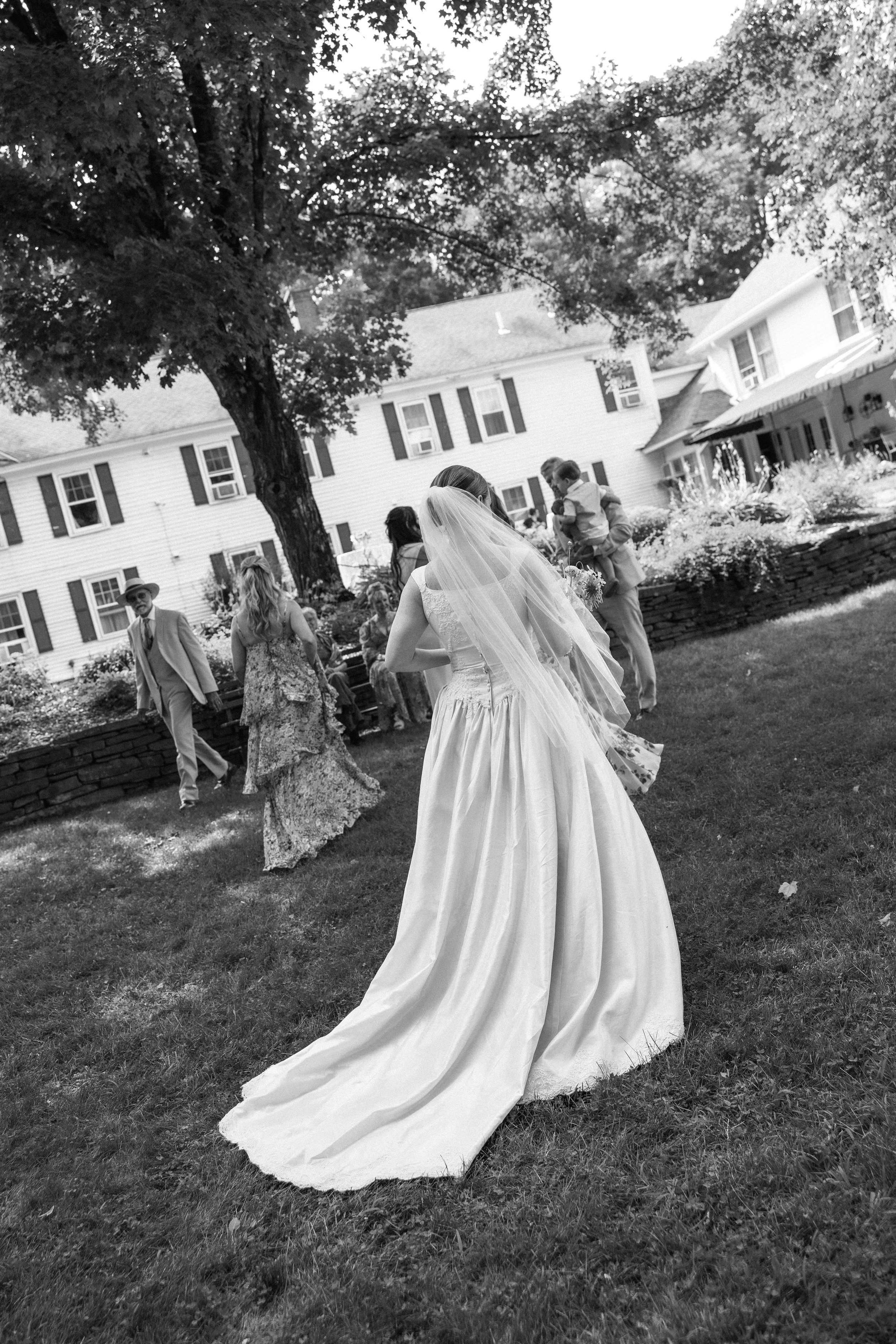 Black and white photo of a bride in a wedding dress with a long train, walking across a grassy yard during a outdoor wedding ceremony, with guests in the background, large trees, and a white house.