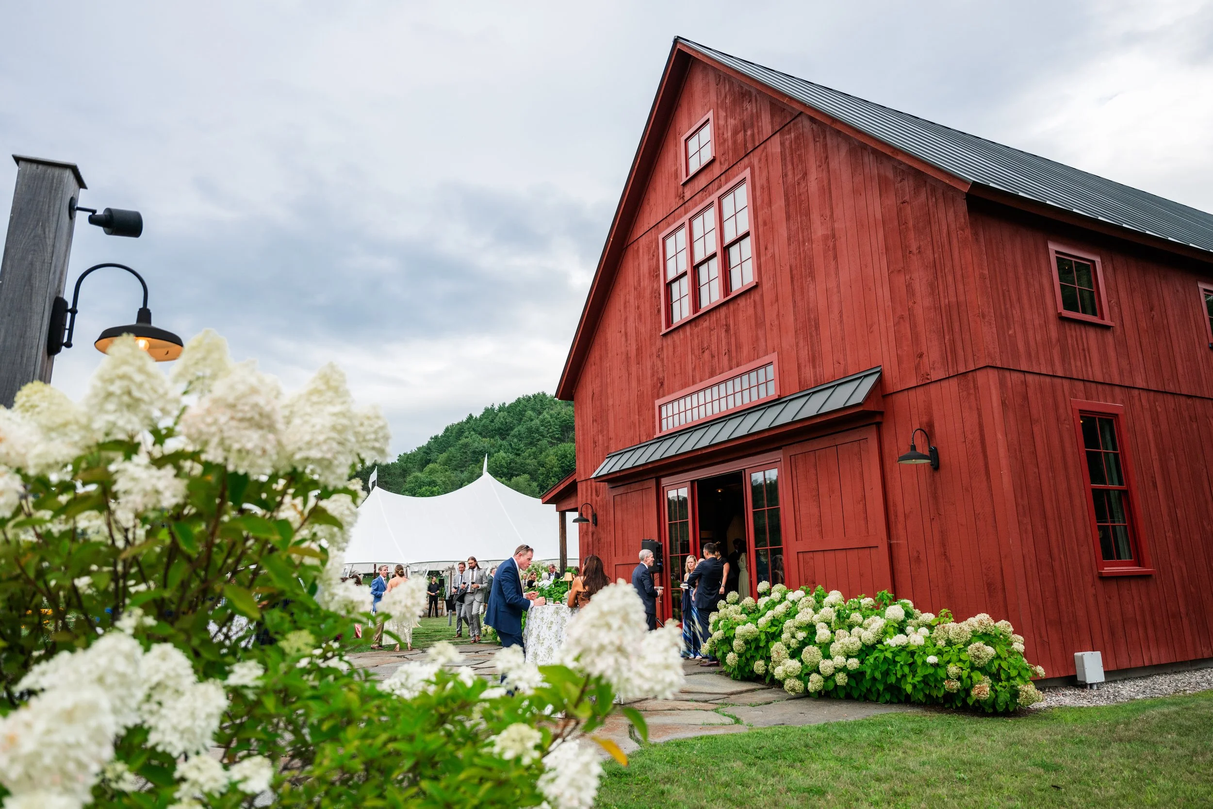 A red barn at an outdoor event with people gathered around tables, surrounded by white flowers and greenery under an overcast sky.