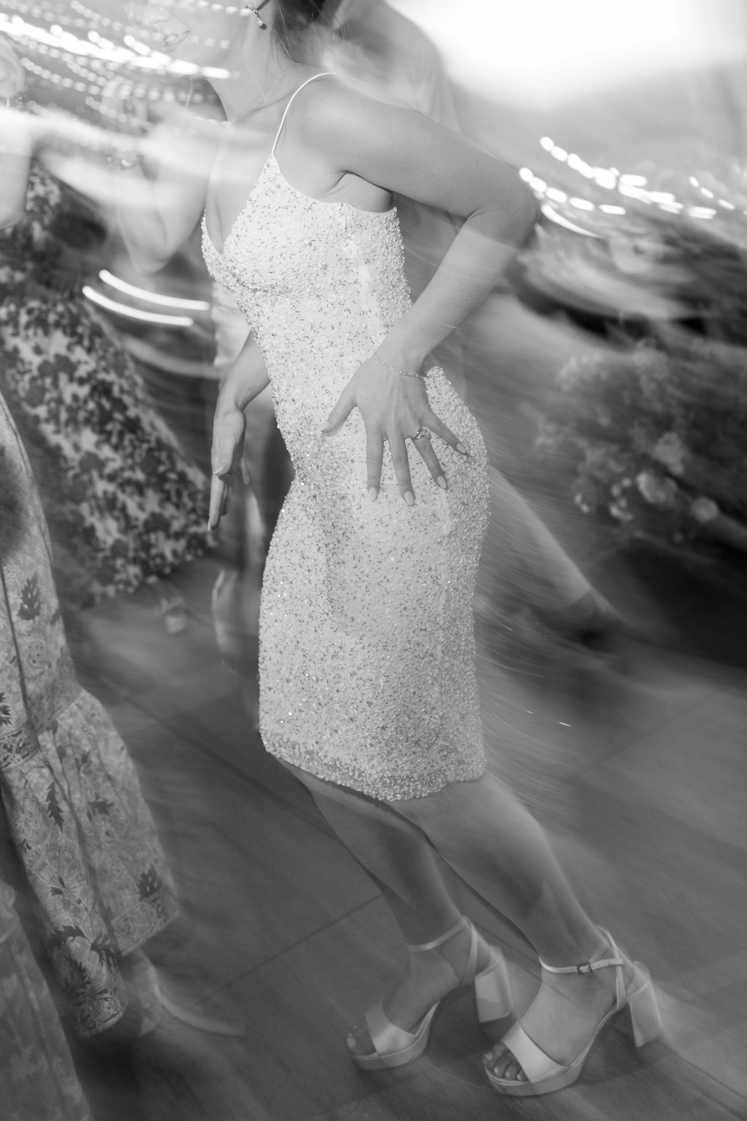 A bride in a jeweled gown and high heels is standing in front of a mirror, with reflections and blurred background around her. New Hampshire wedding.
