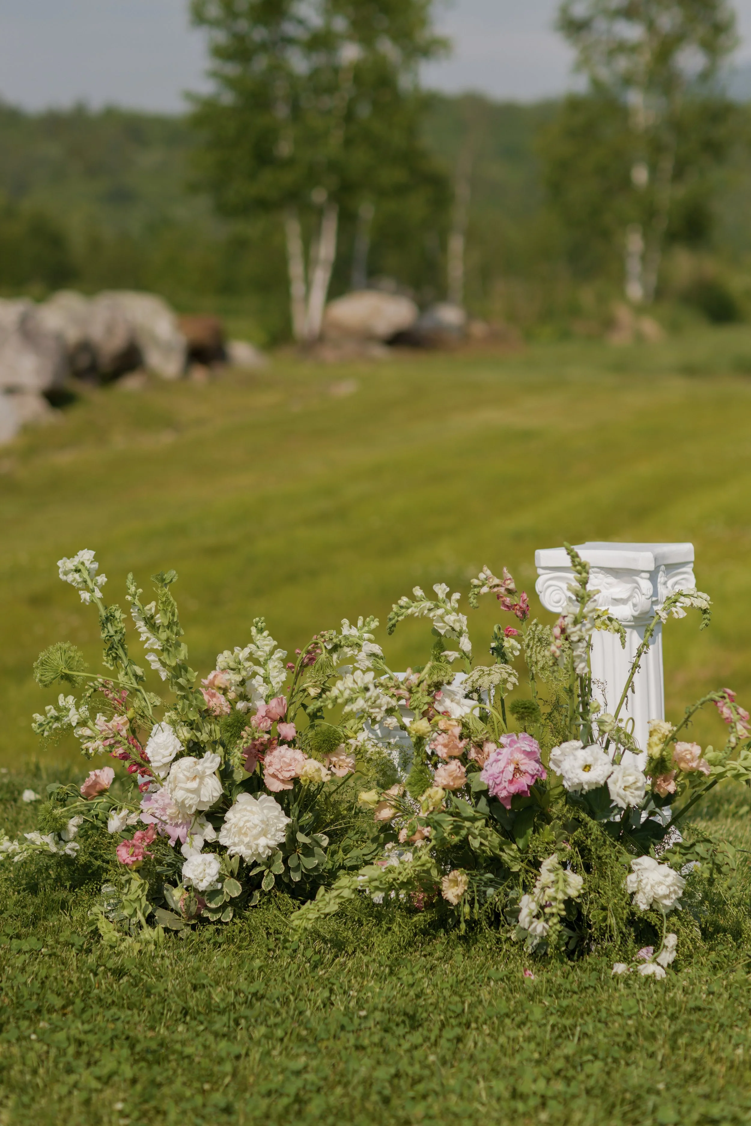 A floral arrangement of pink, white, and green flowers on a white pedestal outdoors, with a grassy field, trees, and rocks in the background. New Hampshire Wedding.