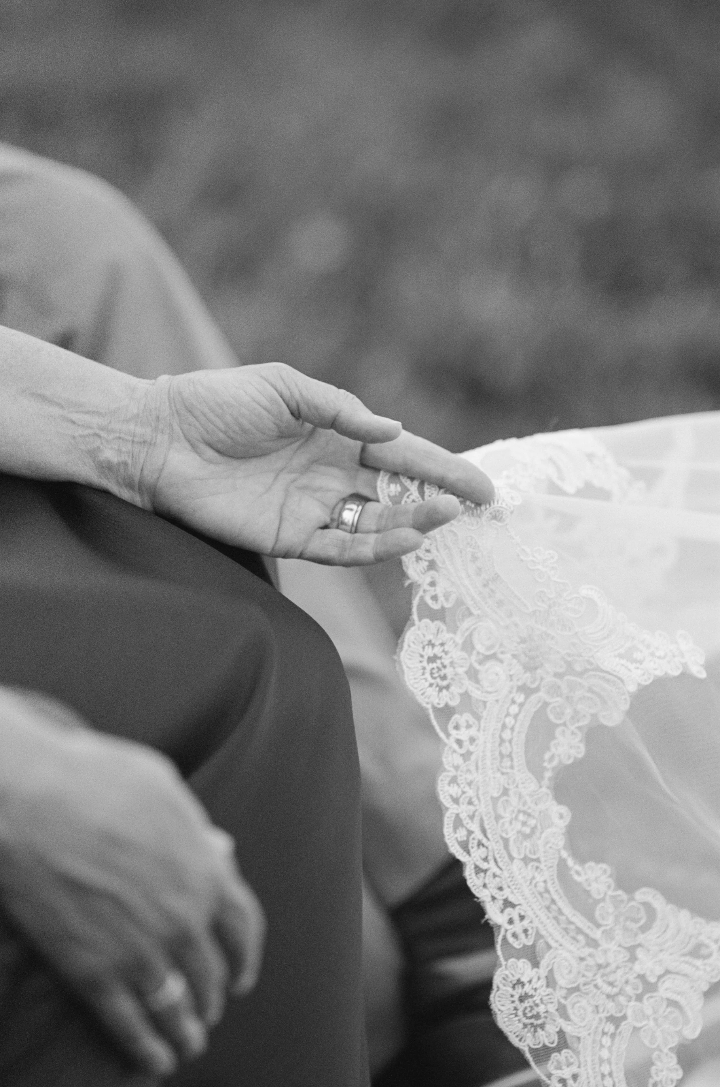 Close-up of a person's hand with a wedding ring, resting on another person's lap, with a lace dress visible. New Hampshire Wedding.