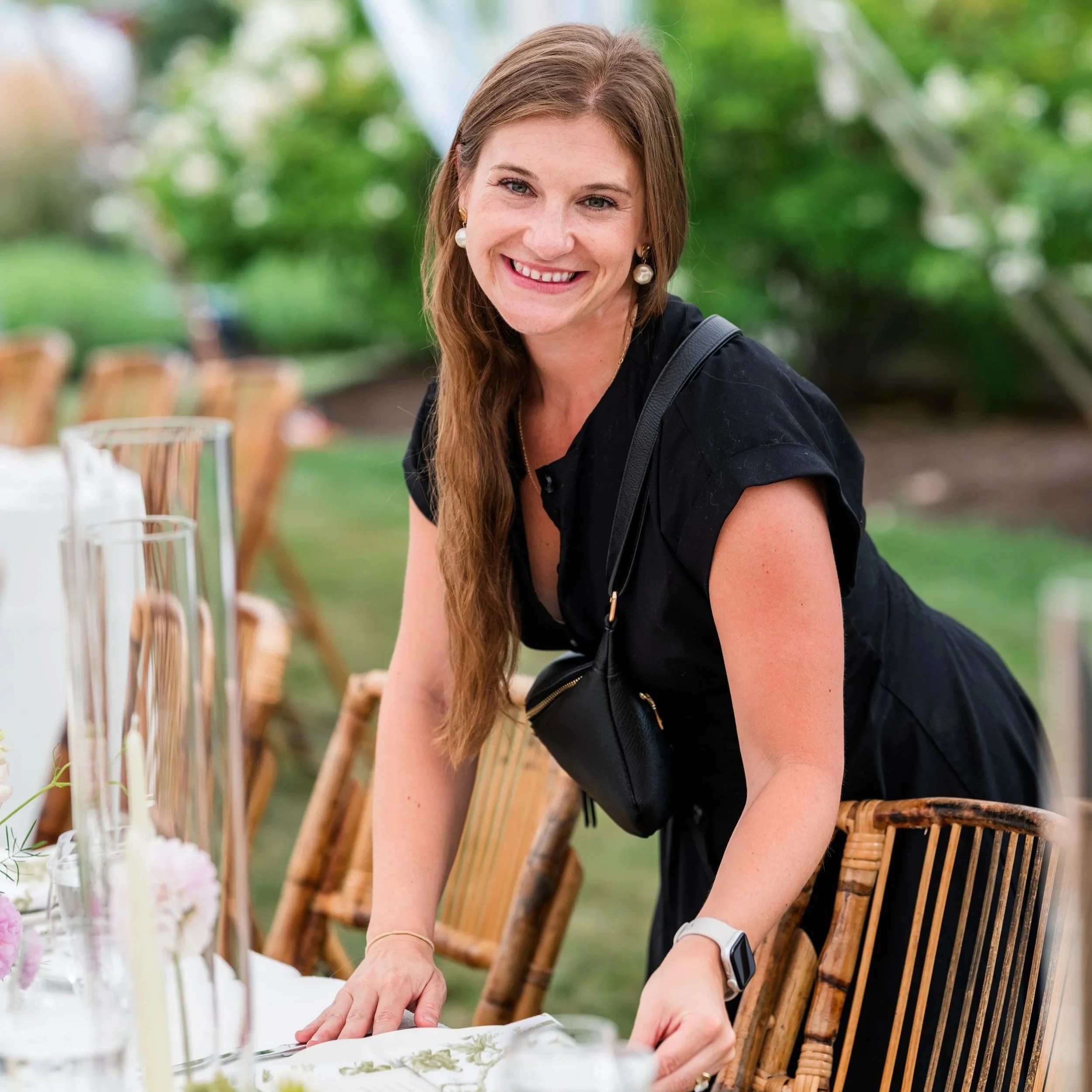A woman with long brown hair, earrings, and a black shirt sitting at a table outdoors, smiling while reaching for food during a gathering with bamboo chairs and greenery in the background.
