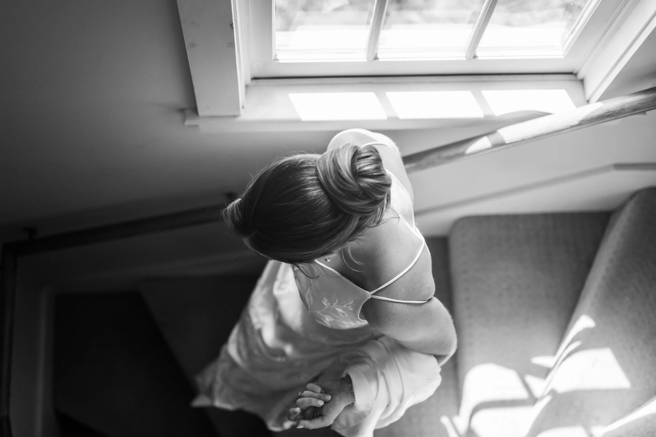 A woman with styled hair and earrings sitting by a window, looking downward, with sunlight coming through the window.