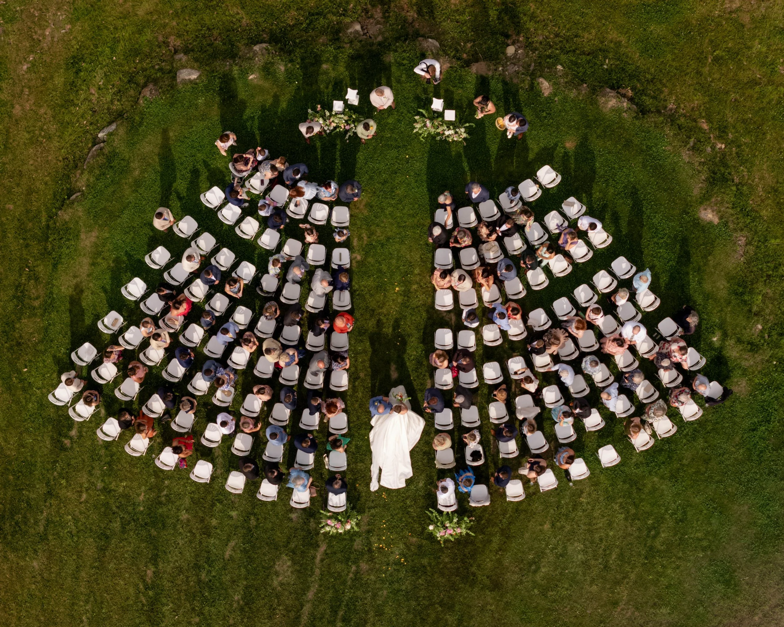 An aerial view of a group of people gathered outdoors for a wedding ceremony on a grassy area, with white chairs arranged in rows and floral decorations. New Hampshire wedding.