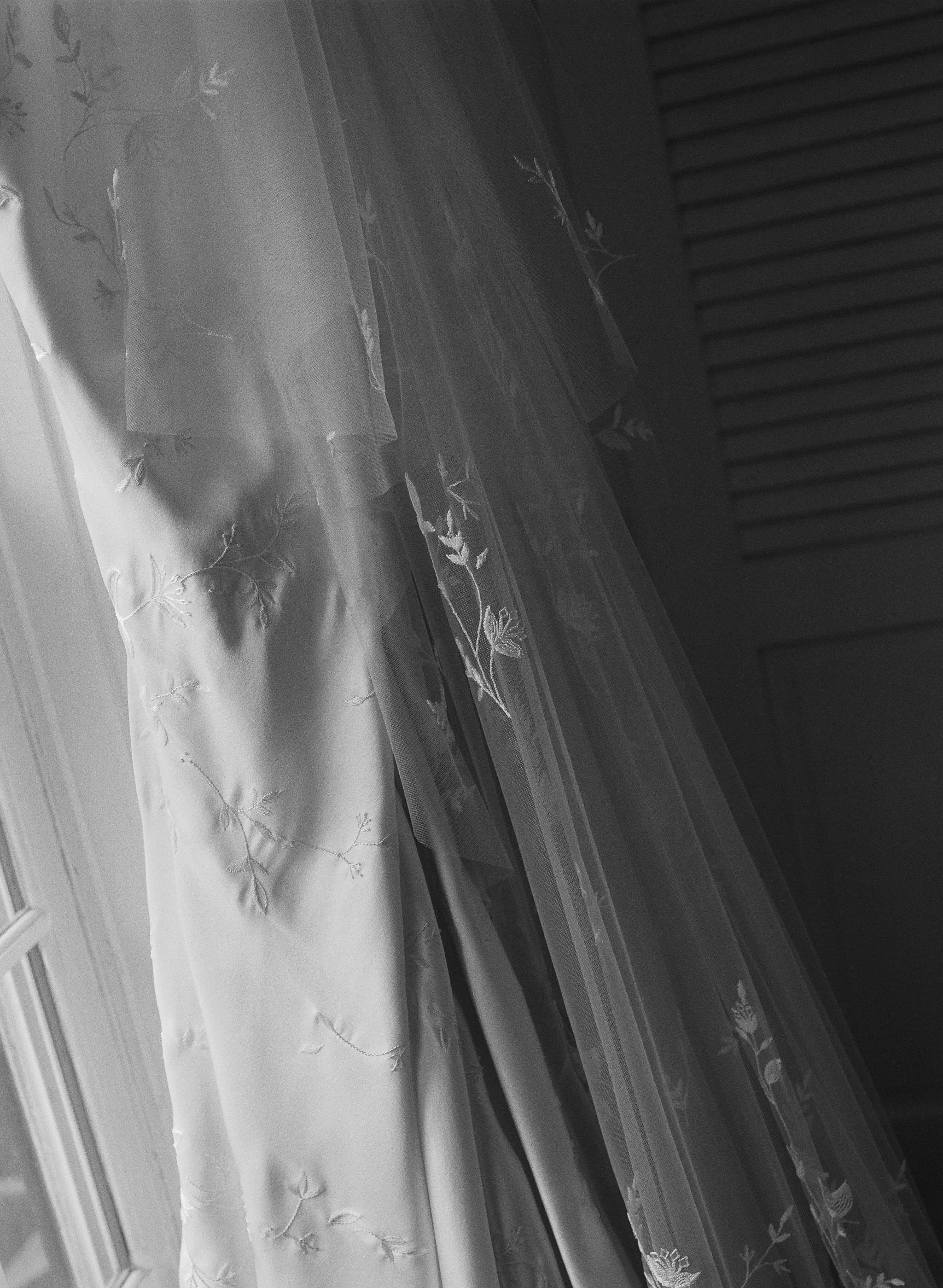 This is a grayscale photo of sheer curtains with floral embroidery hanging in front of a window, partially obscuring the view outside.