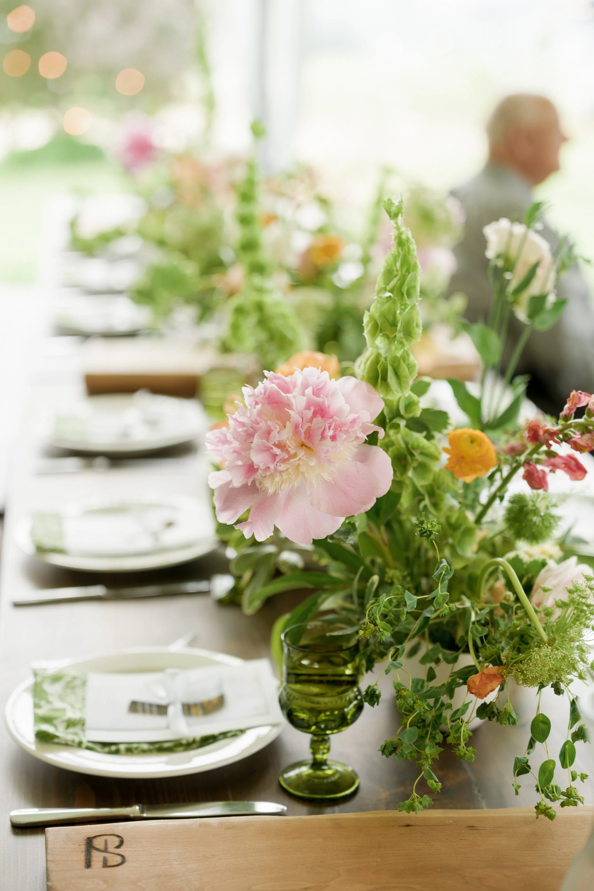 A dining table with a large floral centerpiece in a green glass vase, surrounded by white plates and cutlery, with a blurred background of a person in profile and window light.