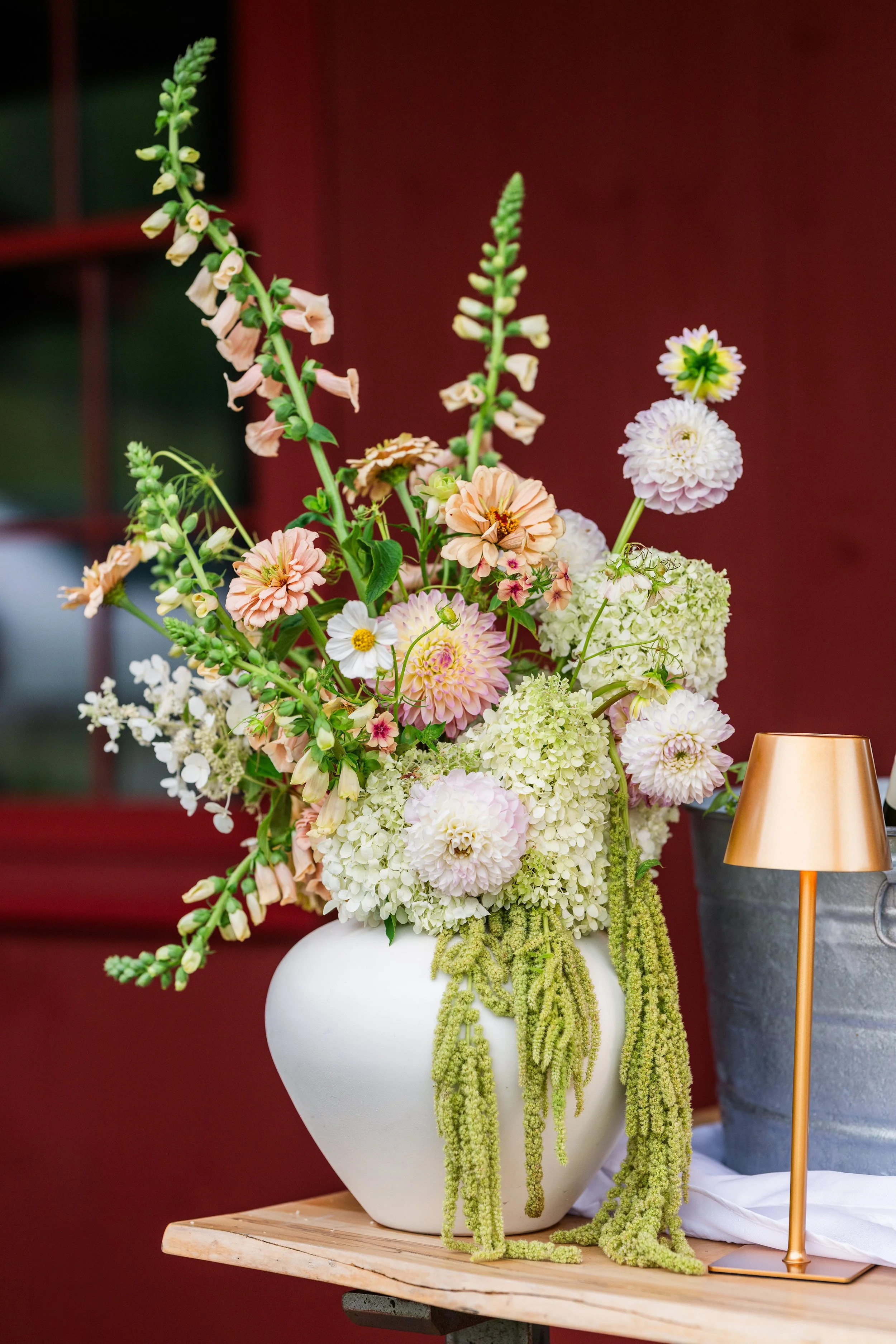 A large white vase containing a colorful flower arrangement on a wooden table, next to a gold-colored table lamp with a gray base.