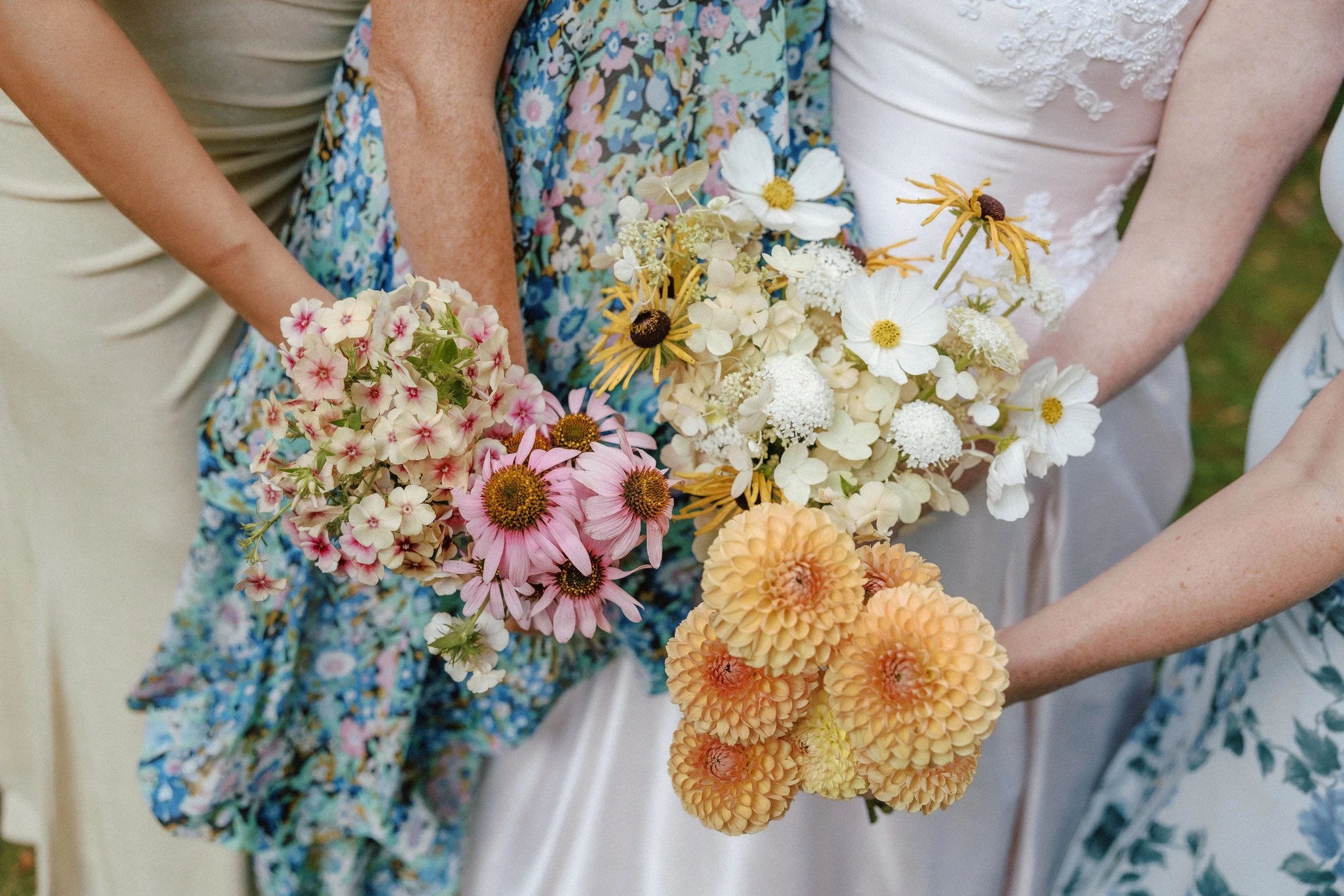 Close-up of women holding colorful flowers at a garden or outdoor event, with floral dresses and garden greenery visible.