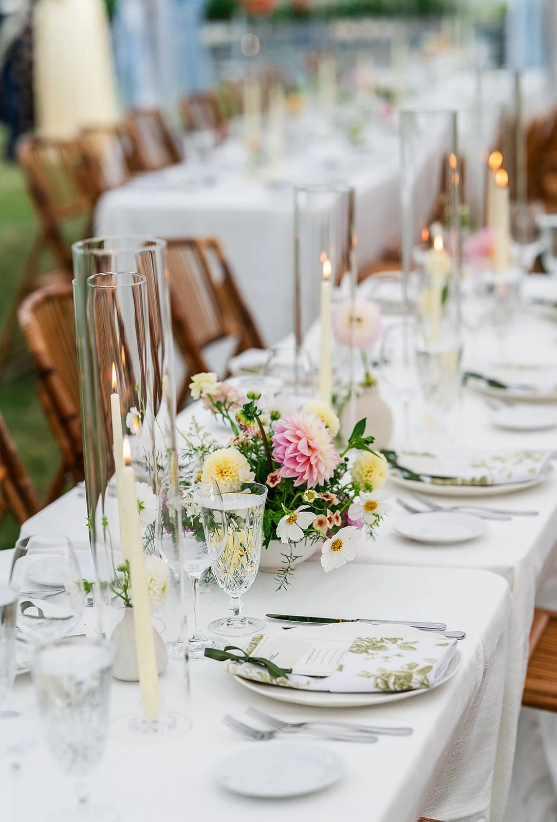 A long dining table decorated with a white tablecloth, floral centerpieces, taper candles in glass holders, elegant glassware, and table settings with plates, napkins, and silverware. Wooden chairs surround the table, and the setting appears to be outdoors or in a bright, airy space.