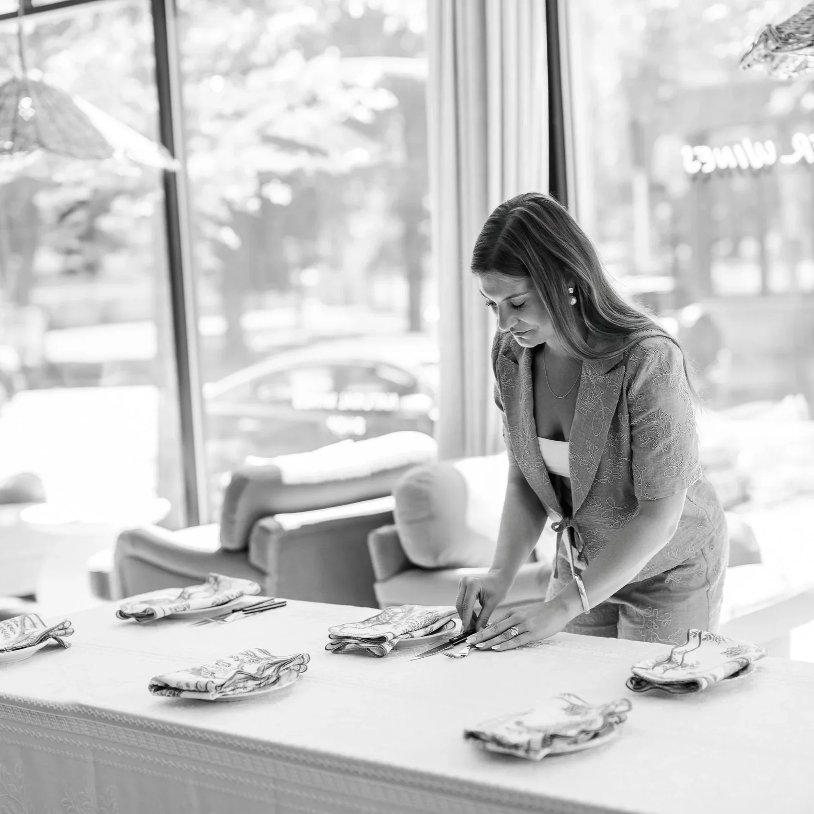A woman is folding napkins at a table in a brightly lit restaurant or cafe, with large windows in the background showing trees and cars outside.