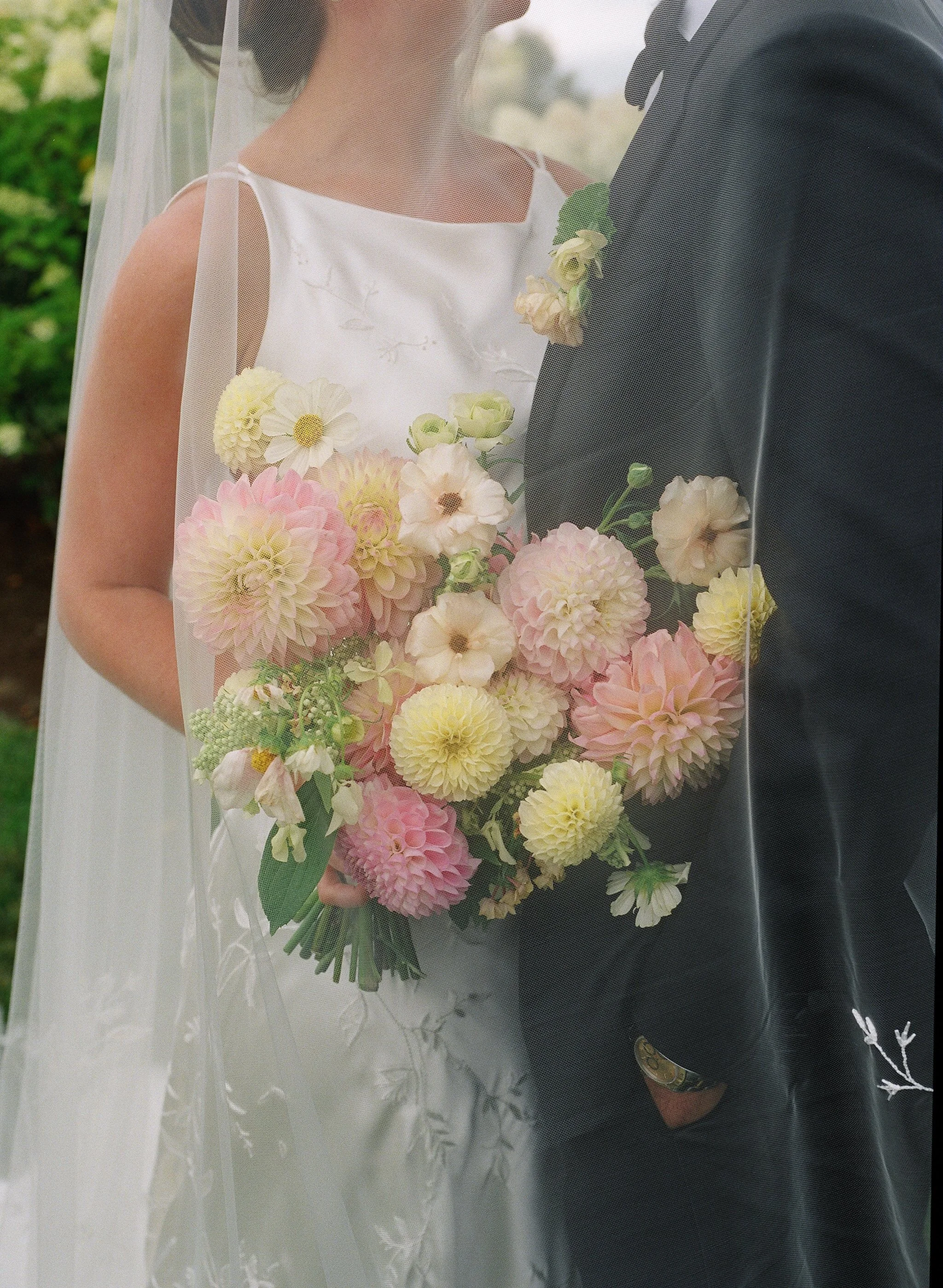 Close-up of a bride and groom standing closely together. The bride is holding a bouquet of pastel-colored flowers, including white, pink, and yellow blooms. The bride wears a white dress with spaghetti straps and a veil, and the groom wears a dark su