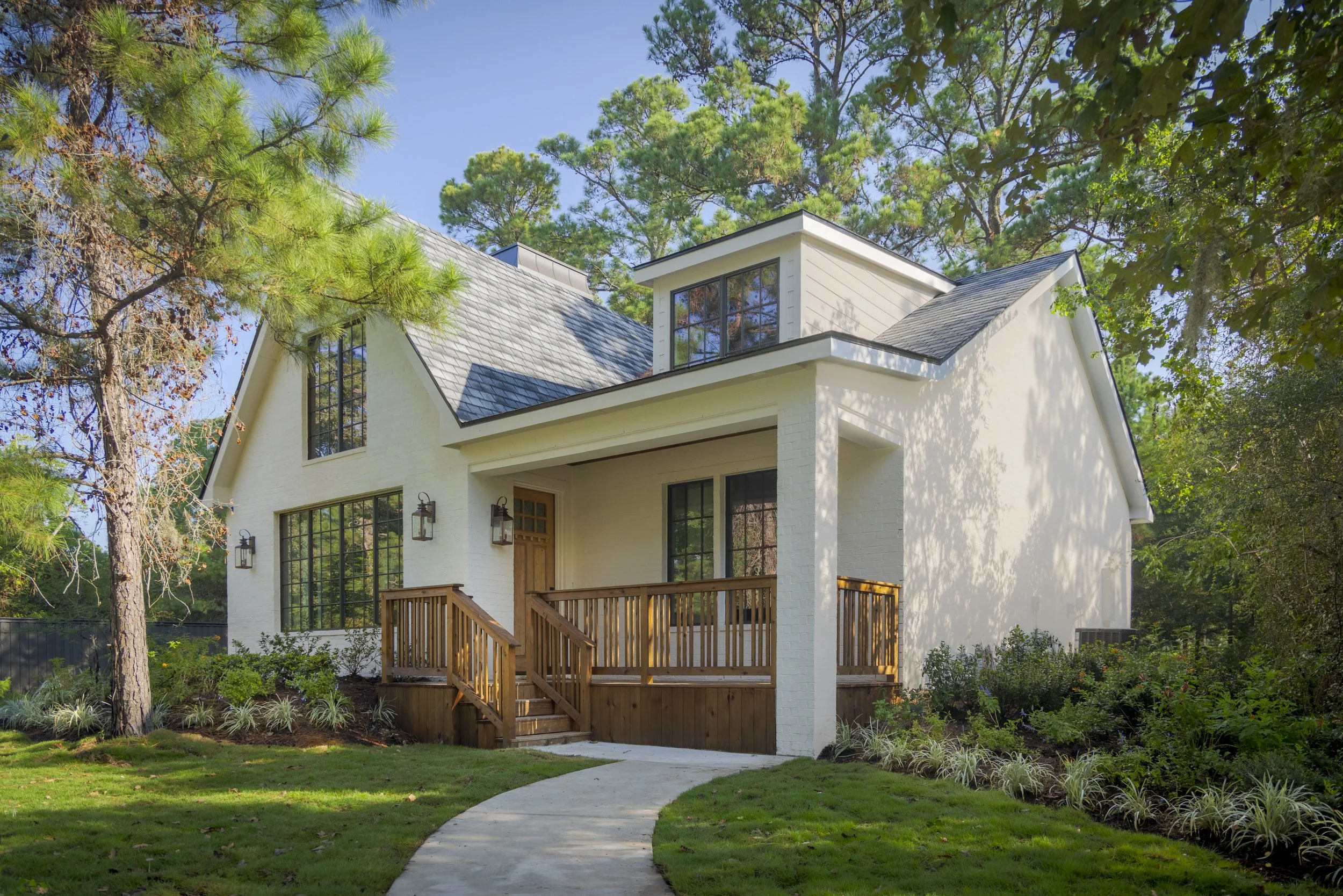 Front view of a modern white house with a sloped roof, large windows, a wooden front porch with stairs, surrounded by green trees and a well-maintained lawn under a blue sky.