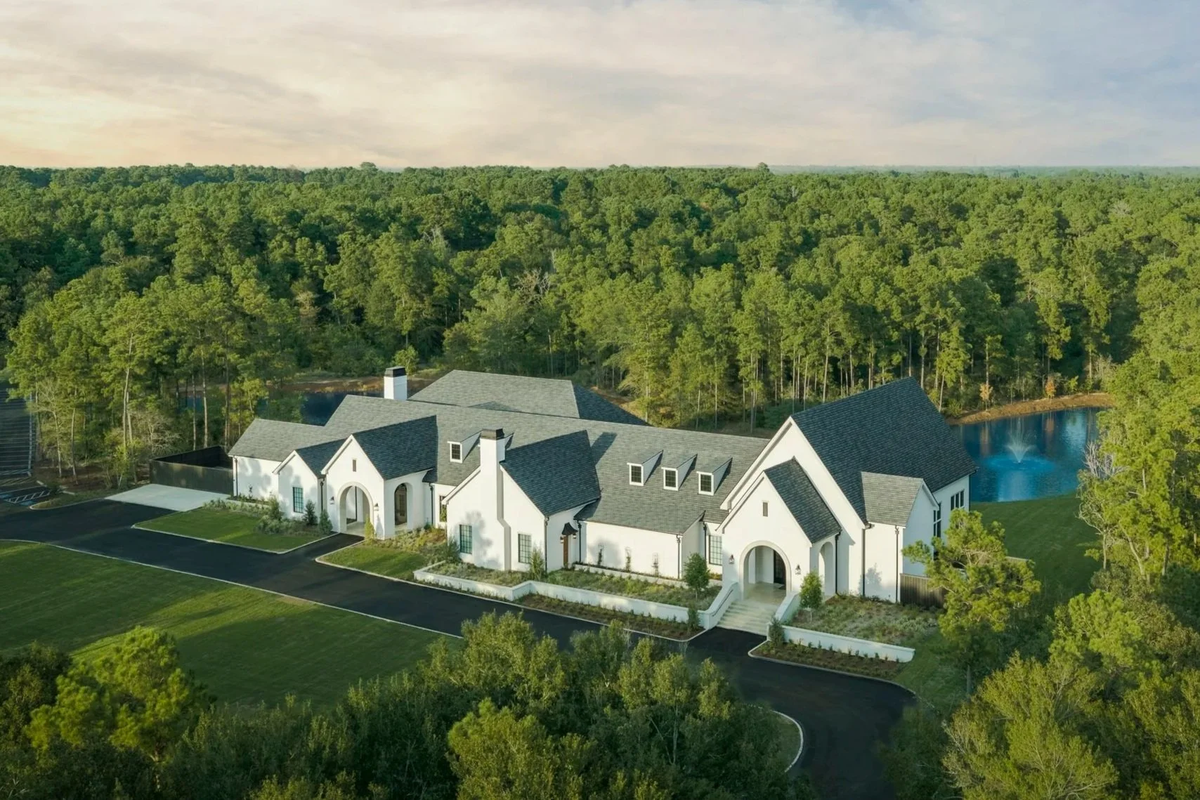 A large white house with a gray roof surrounded by green trees and a landscaped lawn, with a small pond and fountain in the backyard, set near a dense wooded forest.
