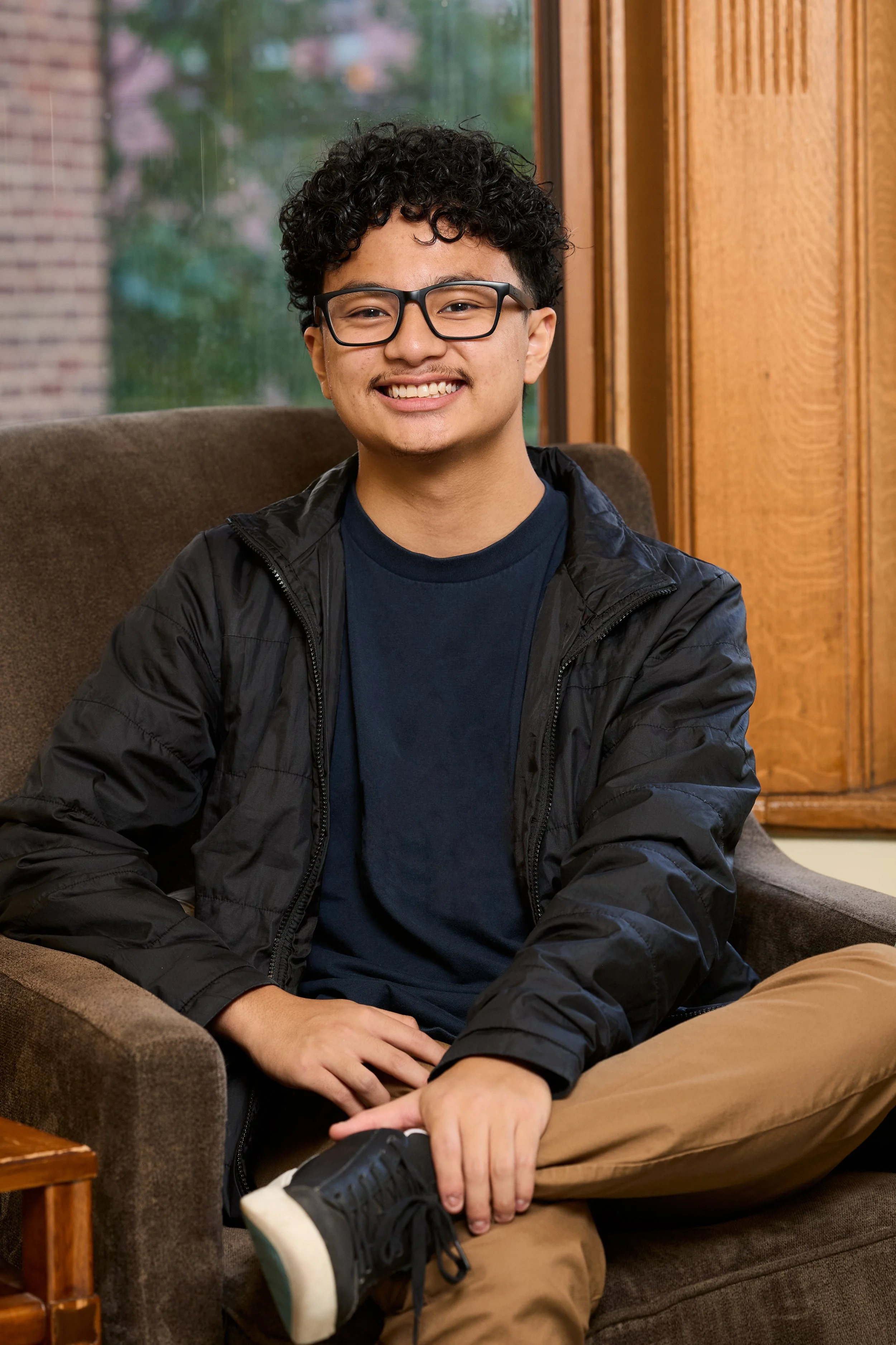 A young man sitting on a brown sofa, smiling, wearing glasses, a black jacket, navy shirt, and khaki pants, in a cozy indoor setting with wood-paneled walls and a window showing trees outside.