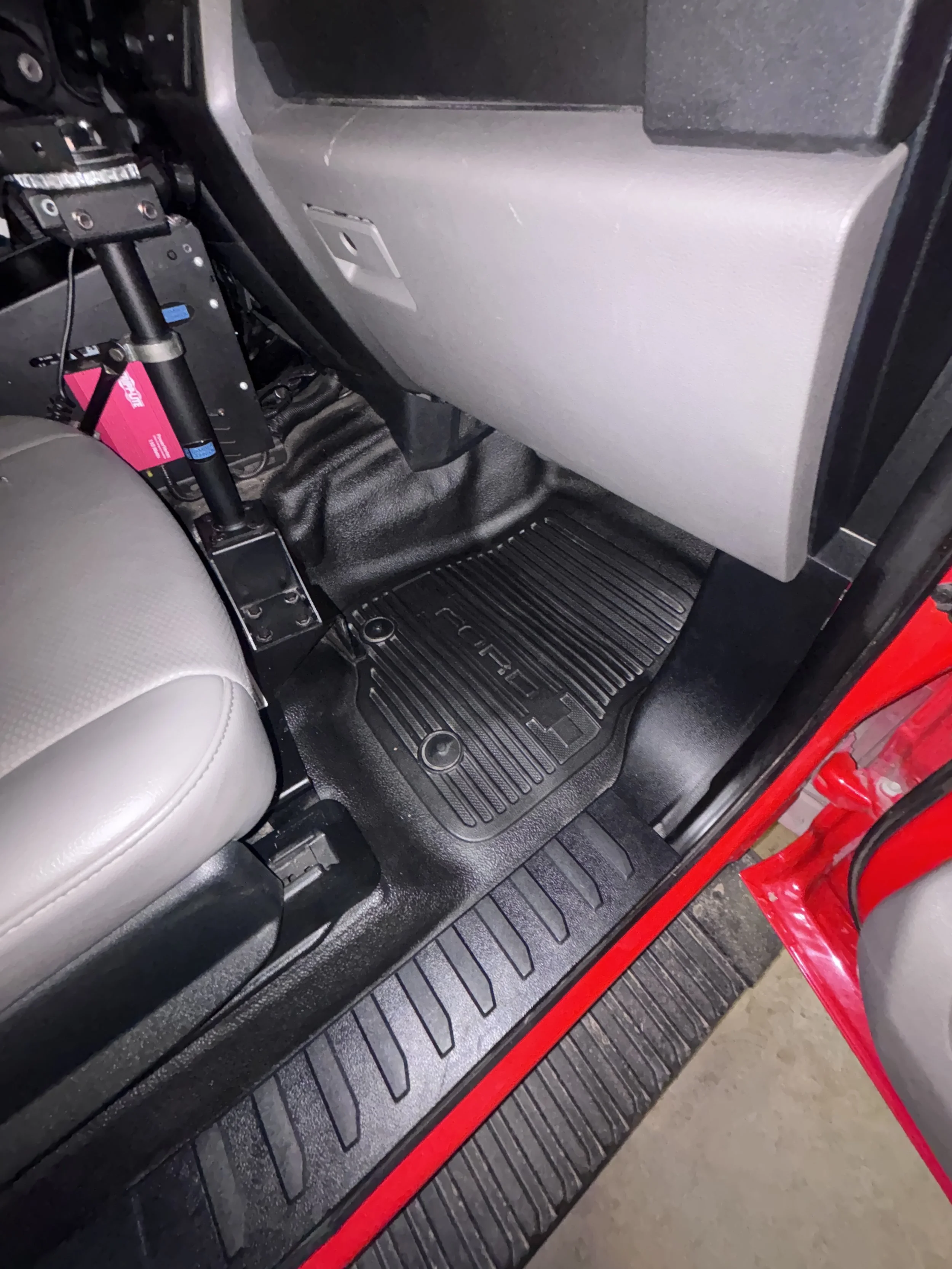 The interior of a vehicle shows the passenger side footwell with a black rubber floor mat, a portion of the beige seat, and a gray glove compartment.