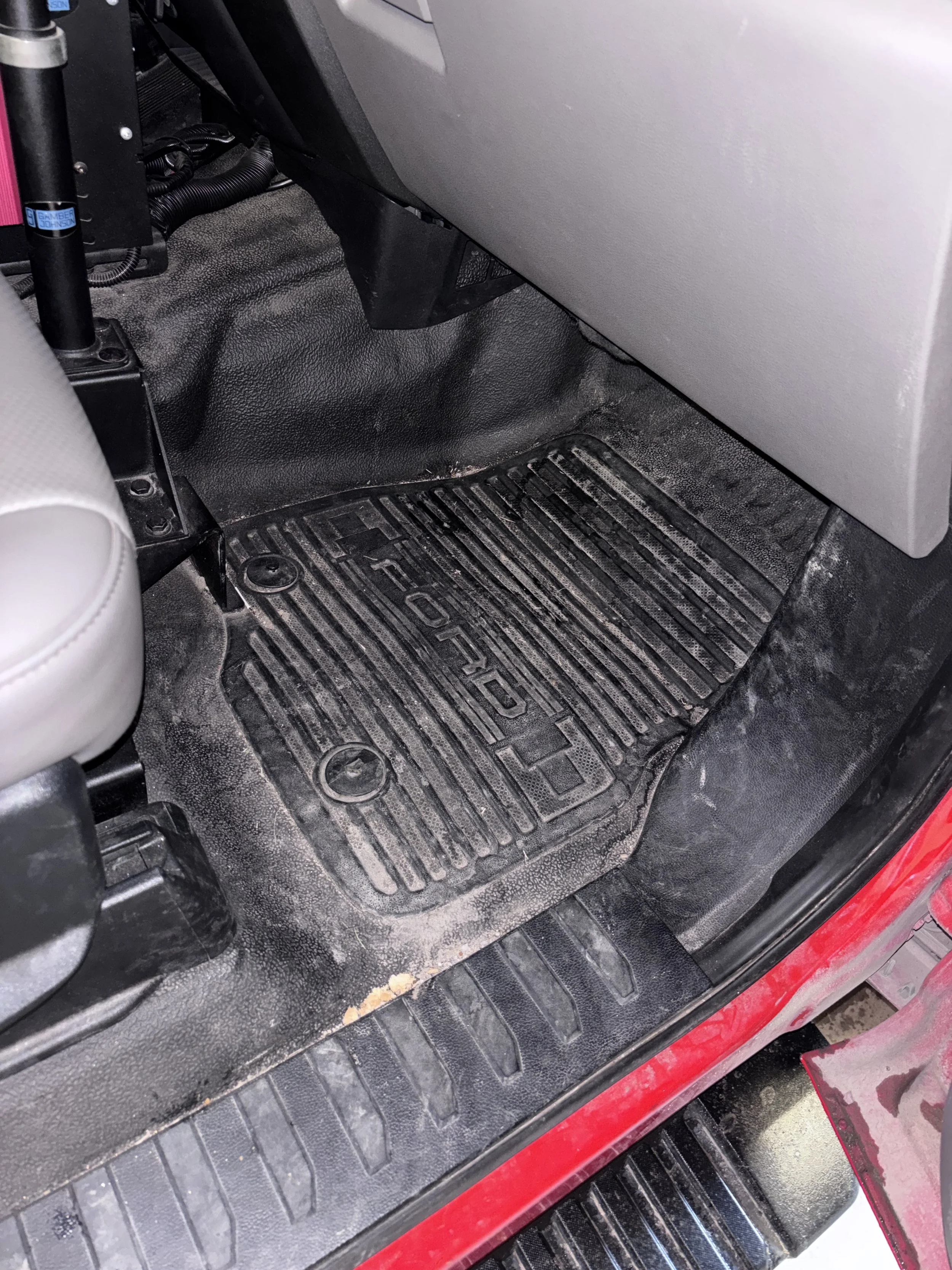 The image shows a dirty rubber floor mat inside a vehicle, with visible dirt and debris. Part of the vehicle's interior, including the edge of a seat and the lower dashboard area, are also visible.