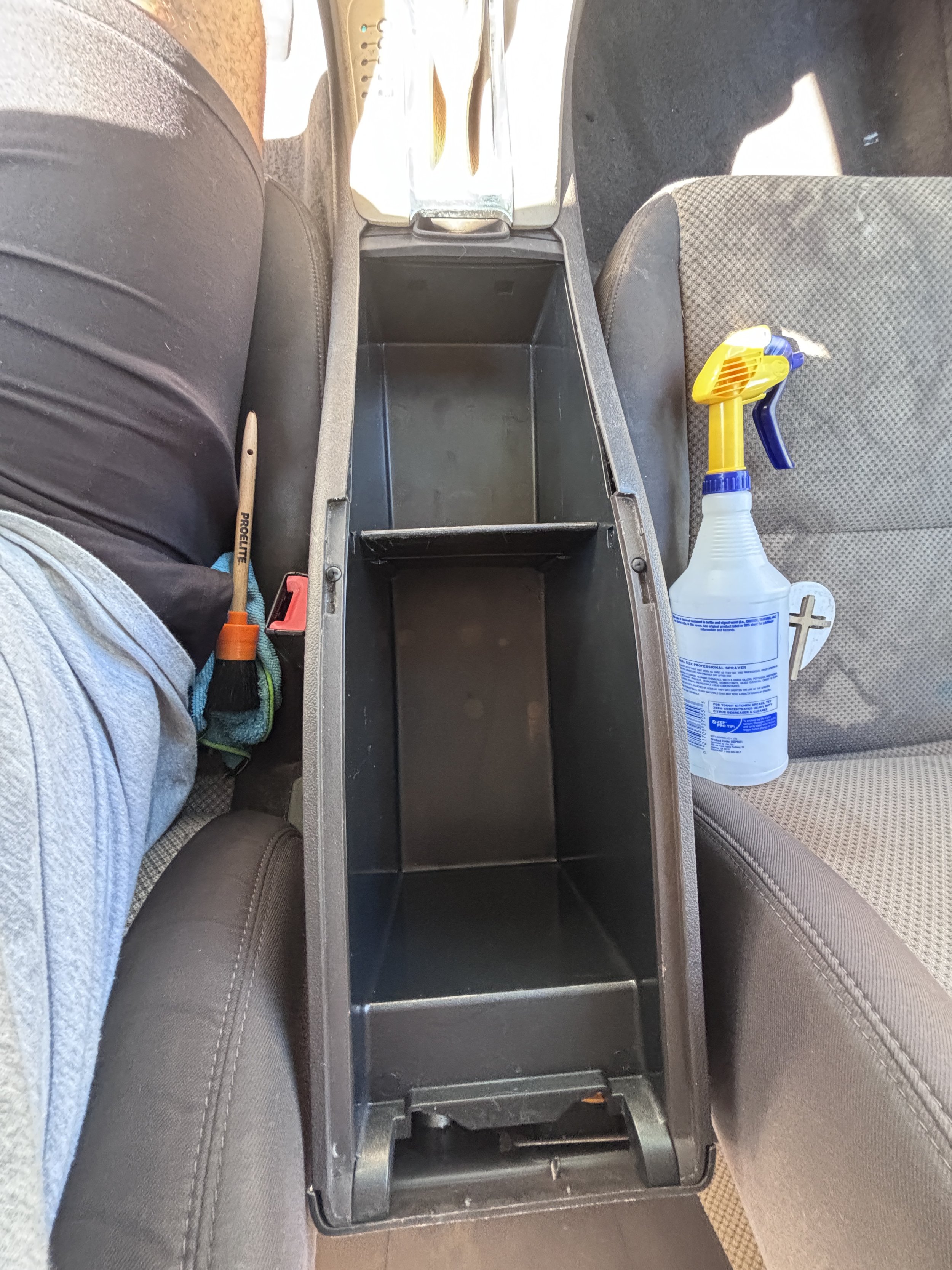 The car's center console with an empty compartment, a spray bottle of cleaner, and a yellow spray nozzle on the passenger seat.