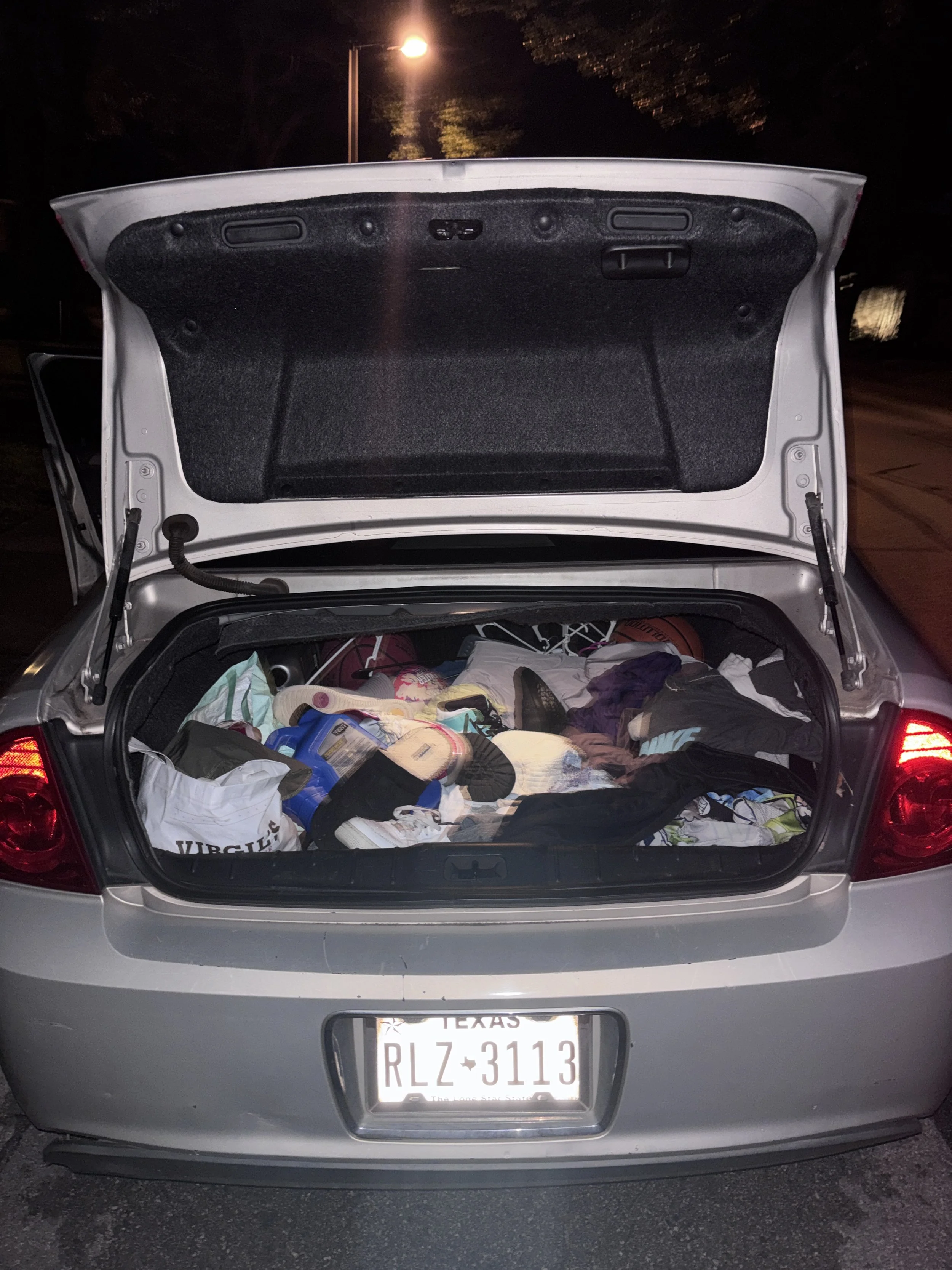 The open trunk of a silver car filled with various items including clothes, bags, and household objects, with a Texas license plate reading RLZ-3113.
