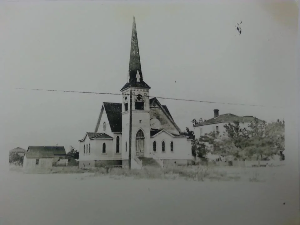 Black and white photo of a church with a tall steeple, surrounded by a few trees and buildings.