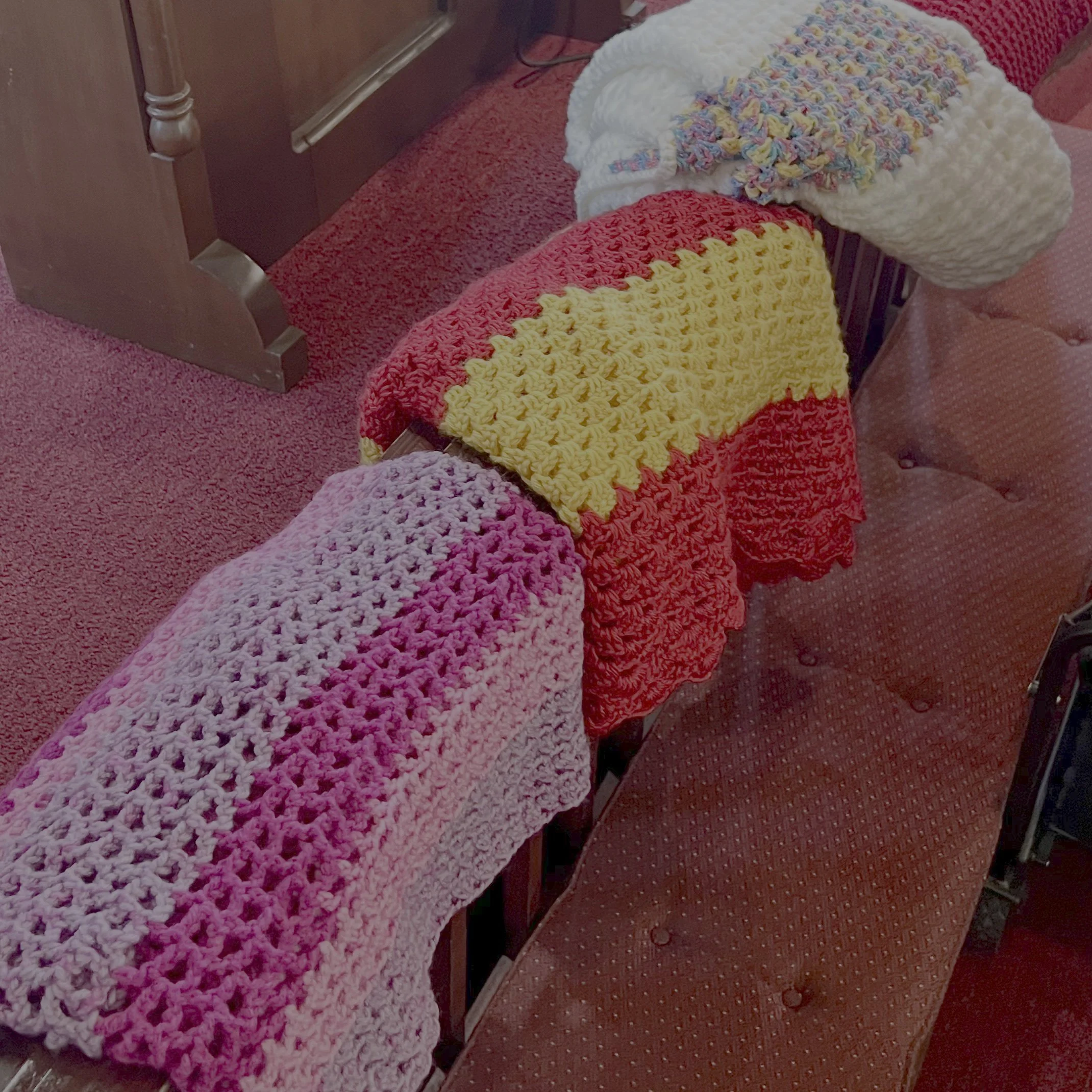 A row of colorful handmade crocheted blankets on a red upholstered bench or bed, with a pink carpeted floor visible on the side.