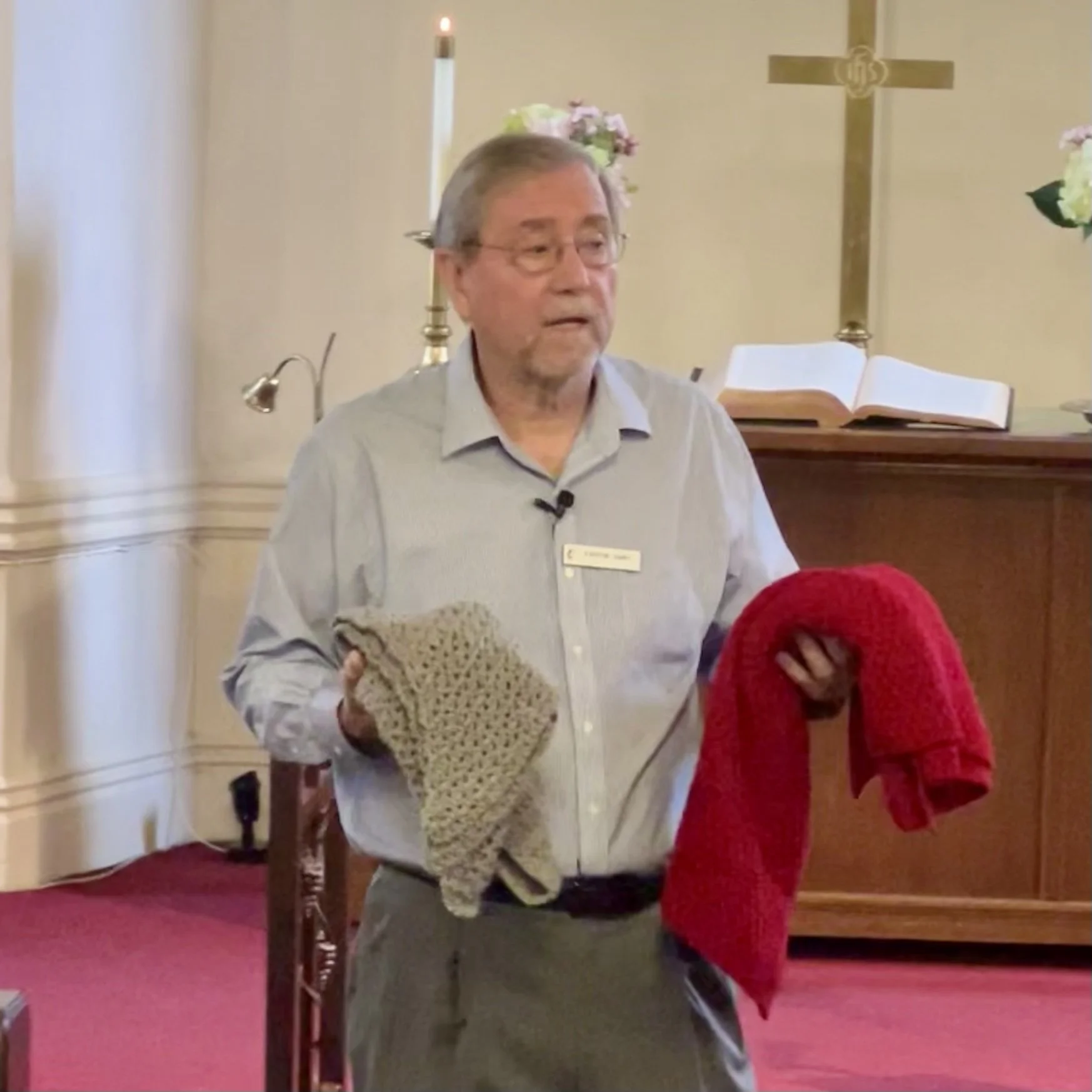Pastor with glasses and a light-colored shirt standing in a church, holding a crochet item