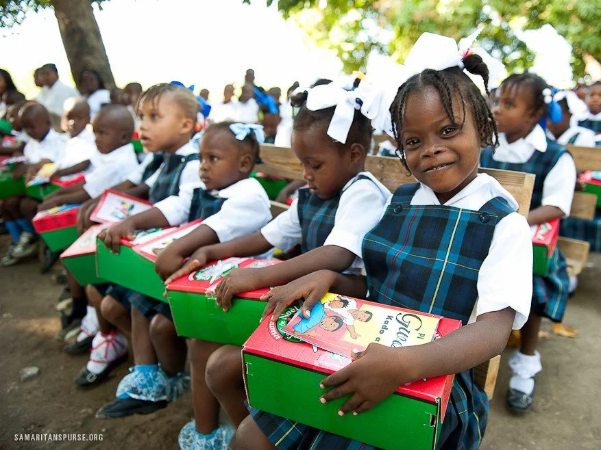 Group of school children in uniform holding and receiving boxes of supplies outdoors during the daytime.
