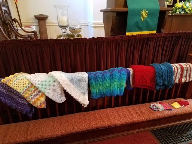 Knitted scarves or shawls hanging on the backrest in a church. In the background, there are candles and a green cloth with gold lettering.