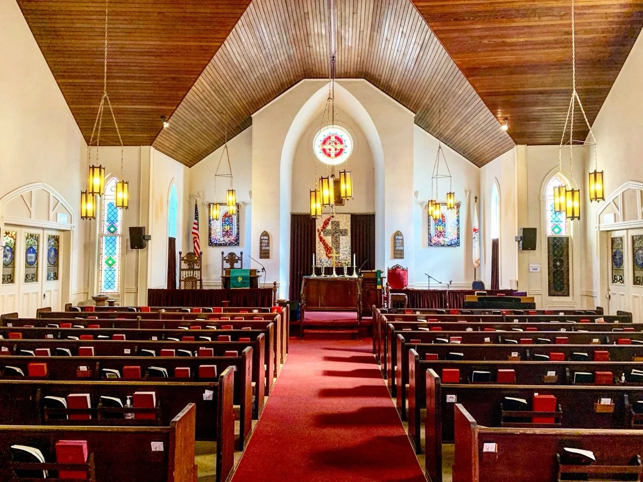 Interior of a church sanctuary decorated for a wedding with wooden pews, stained glass windows, and an altar with a cross, candles, and banners at the front.