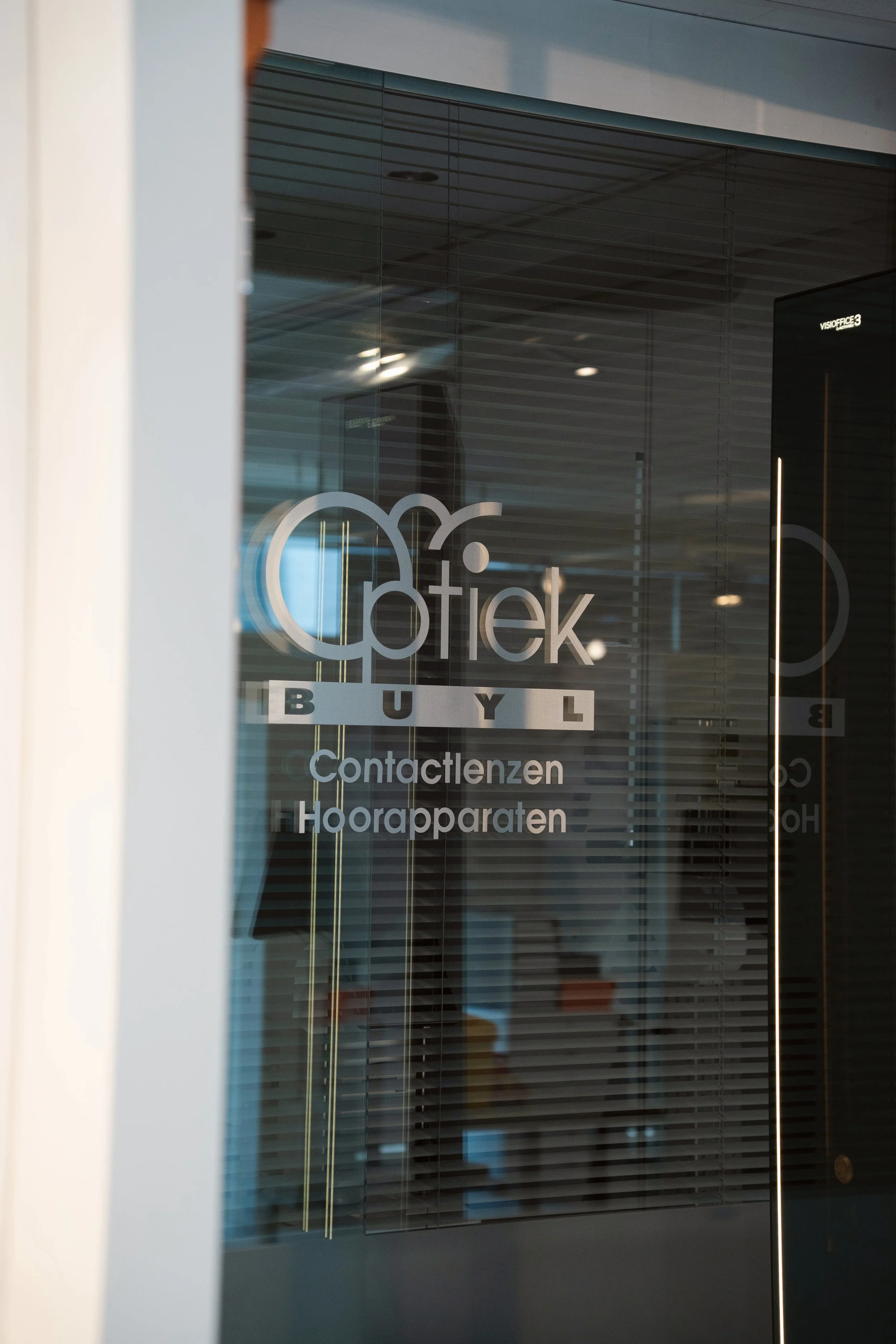 Glazen kantoorwand met het logo van Bitler en de tekst ‘Contactlenzen’ en ‘Hoordapparaten’.