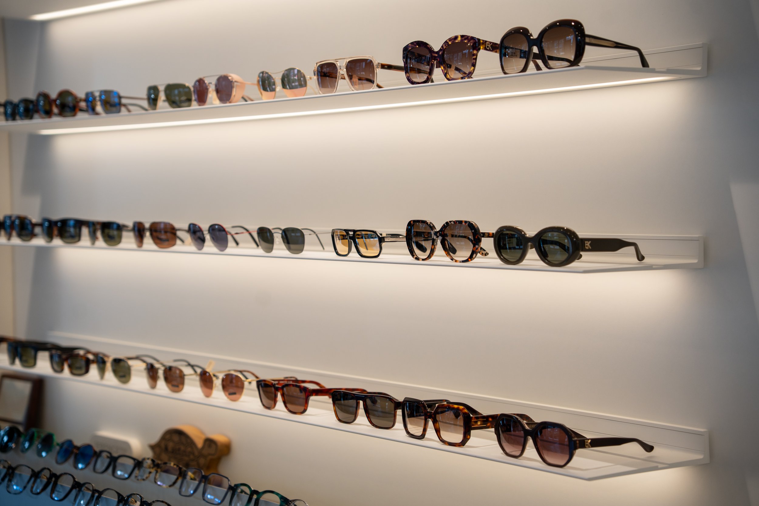 Collectie zonnebrillen en brilmonturen op verlichte wandplanken in een optiekwinkel
